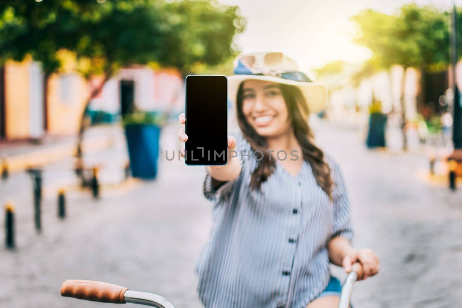 Smiling tourist on bicycle showing cell phone screen on the street. Beautiful girl with hat on bicycle showing cell phone screen on the street by isaiphoto