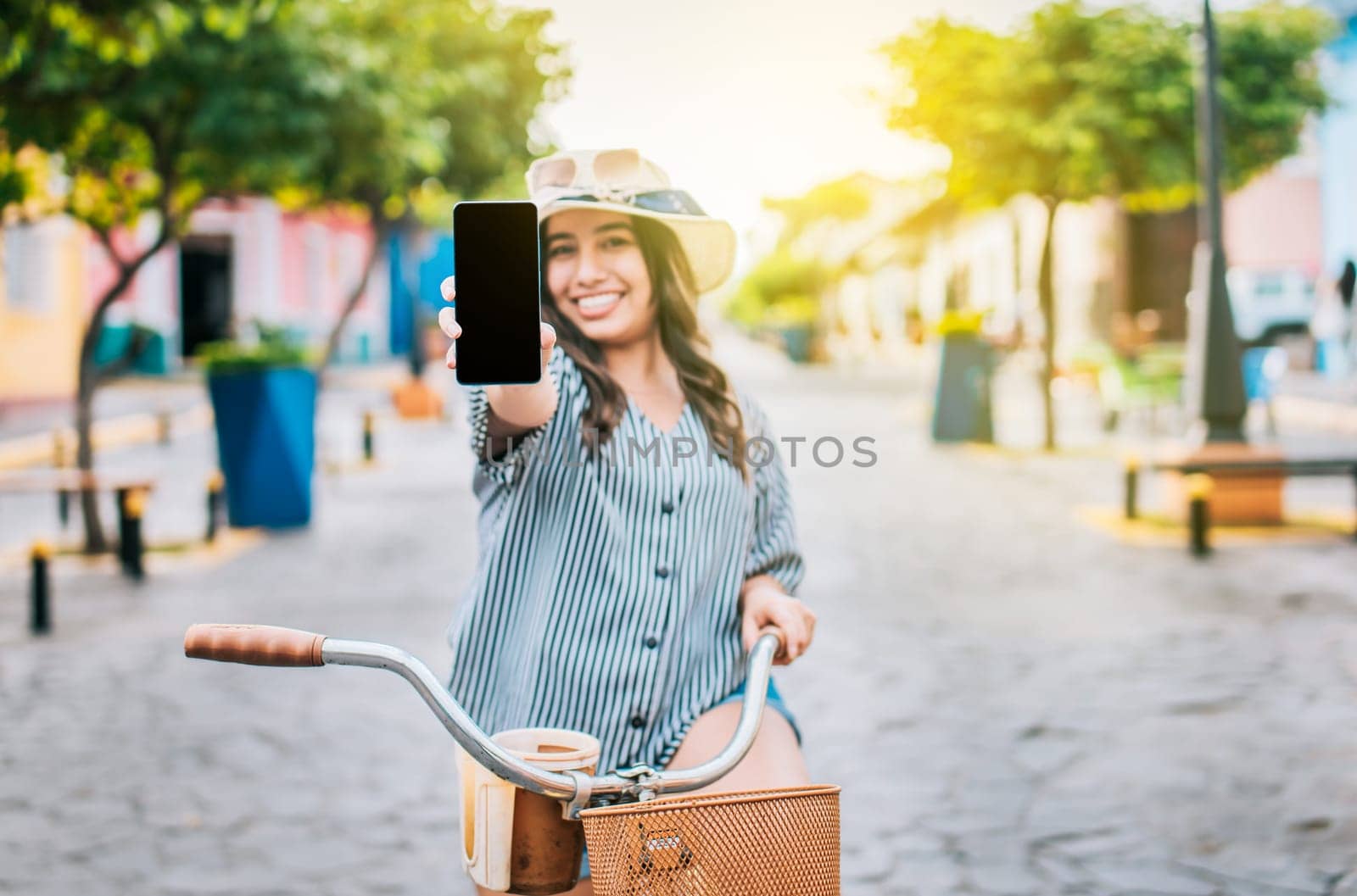Beautiful girl with hat on bicycle showing cell phone screen on the street. Smiling tourist on bicycle showing cell phone screen on the street by isaiphoto