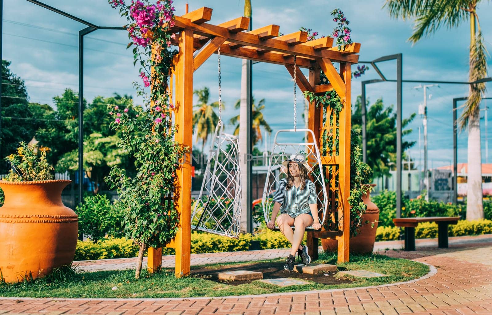 Beautiful girl in hat sitting on a white swing in a beautiful garden. Portrait of a smiling girl sitting on a swing in La Calzada, Granada, Nicaragua by isaiphoto