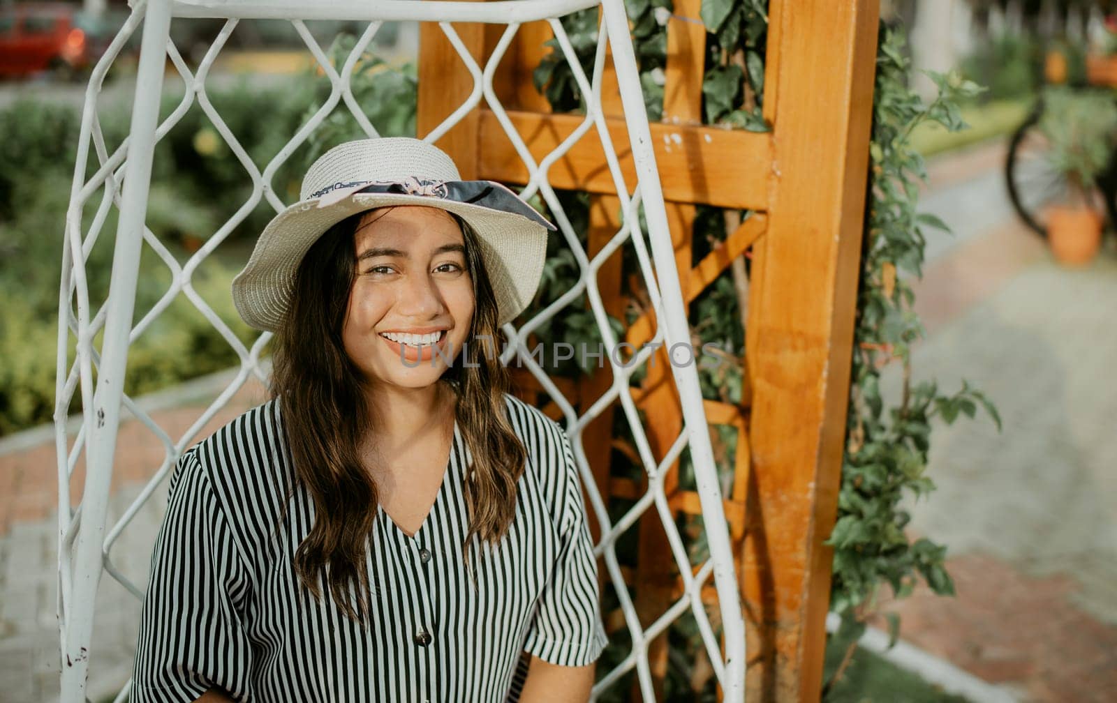 Portrait of a smiling girl in a hat sitting on a white swing in a garden. Lifestyle of girl in hat sitting on a swing looking at camera by isaiphoto