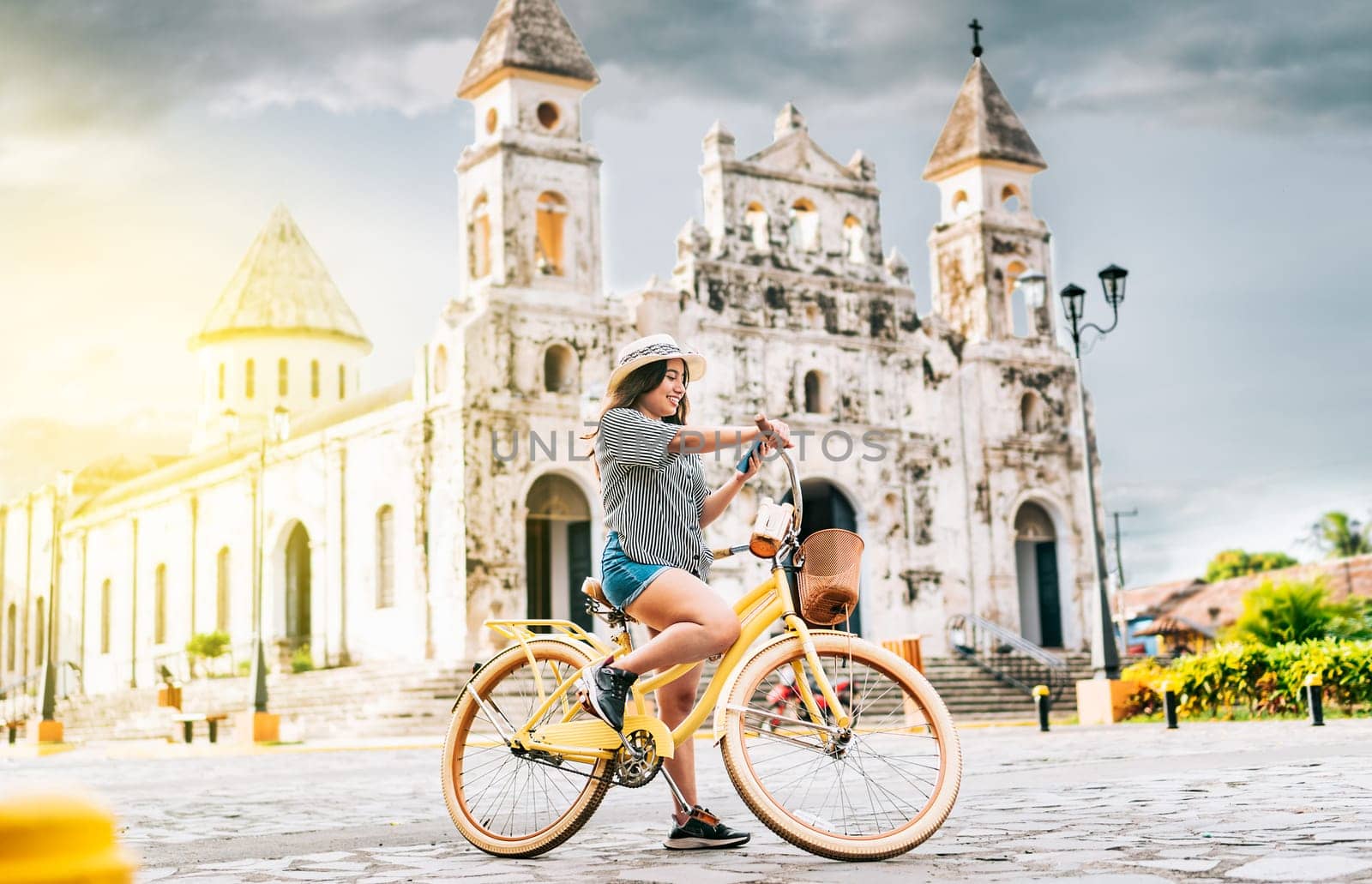 The girl is on the street smiling at the cell phone while she is on her bicycle, there is a church in the background on a beautiful day