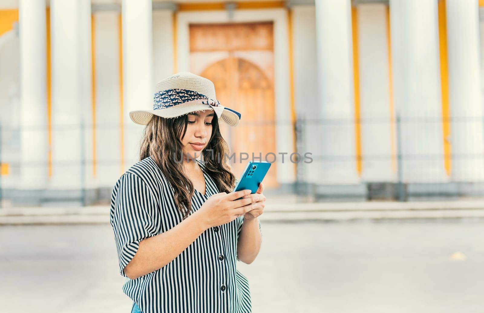 The girl wears a hat, stands in front of a Catholic church with a blurred background, is smiling and sending messages from her cell phone.