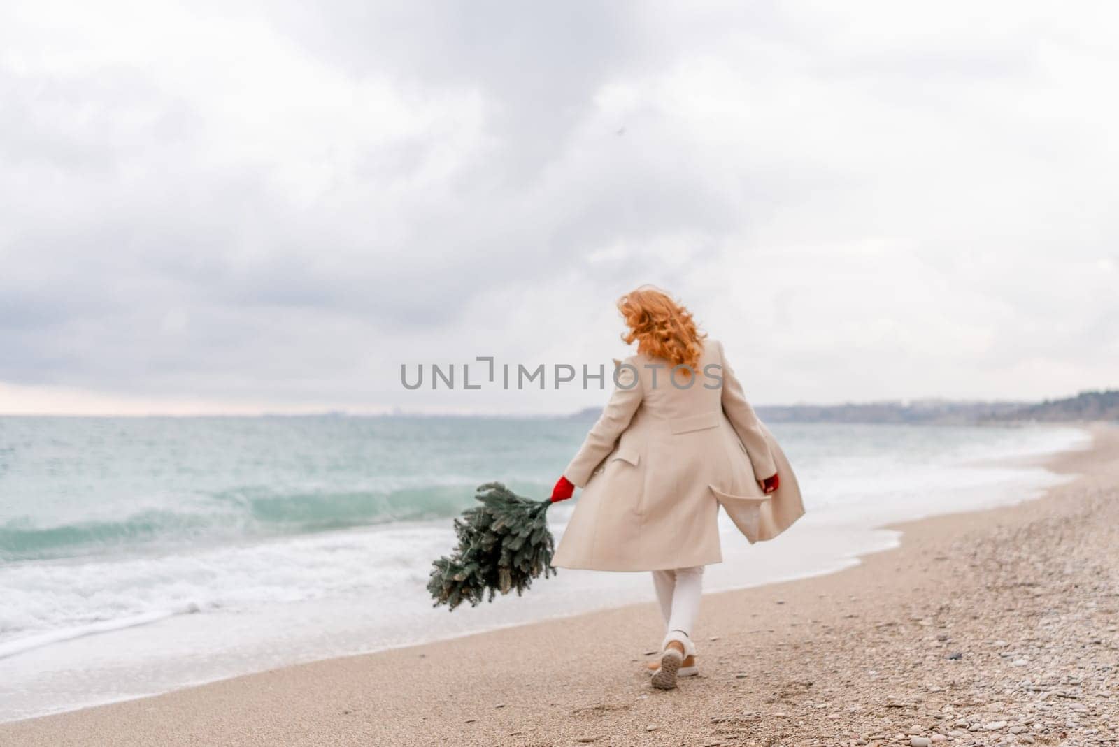Redhead woman Christmas tree sea. Christmas portrait of a happy redhead woman walking along the beach and holding a Christmas tree in her hands. Dressed in a light coat, white suit and red mittens. by Matiunina