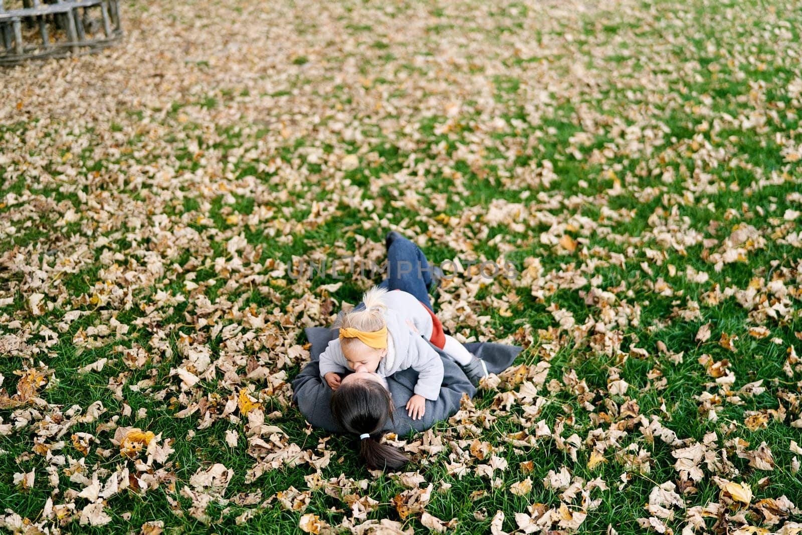 Little girl lies on her mother stomach on the autumn lawn and kisses her on the cheek. High quality photo