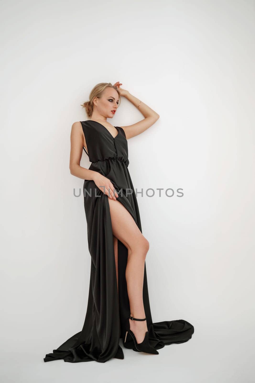 Woman in black dress on white background in the studio. Elegant lady in long gown showing leg. Sexy girl in formal high slit prom dress