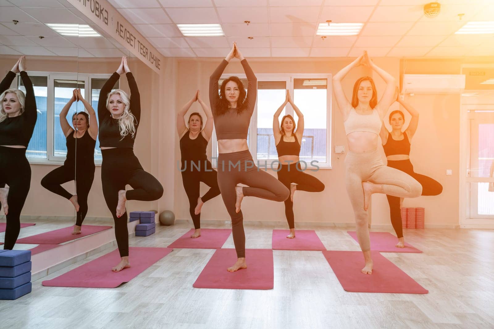 A group of six athletic women doing pilates or yoga on pink mats in front of a window in a beige loft studio interior. Teamwork, good mood and healthy lifestyle concept