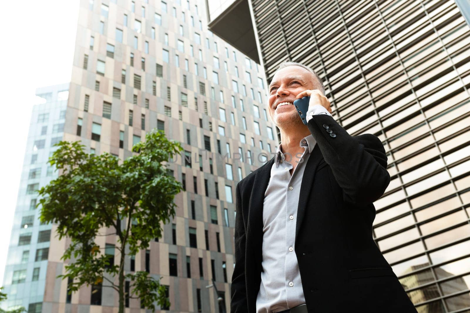 Smiling and successful businessman talking on mobile phone next to office buildings. Copy space. by Hoverstock