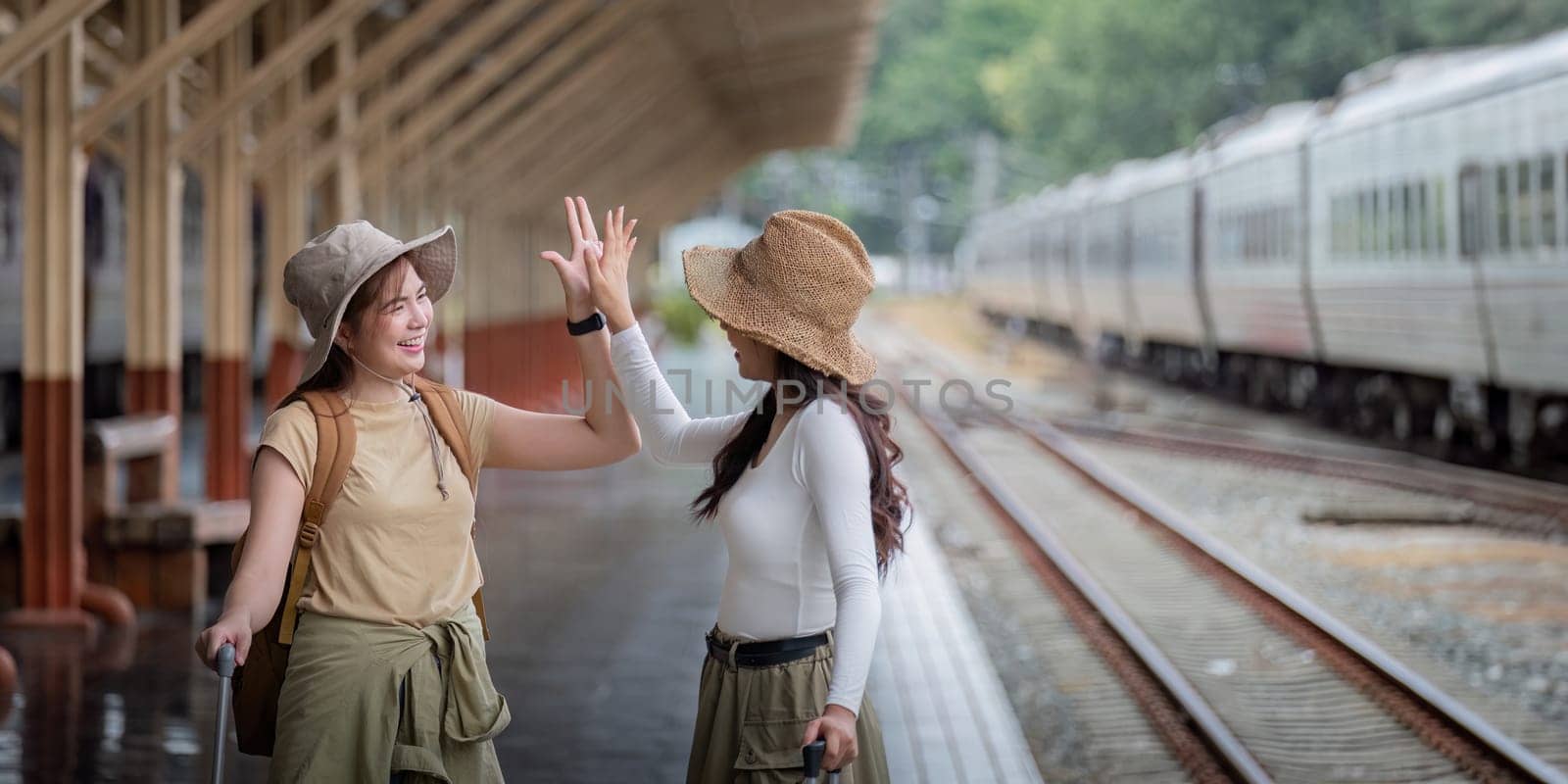 Two women are happy while traveling at the train station. tour concept. Two asian women best friend travel high five on train timetable in railway station by nateemee