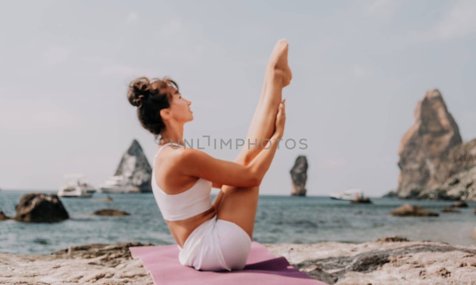 Fitness woman sea. A happy middle aged woman in white sportswear exercises morning outdoors on a beach with volcanic rocks by the sea. Female fitness pilates yoga routine concept. Healthy lifestyle. by panophotograph