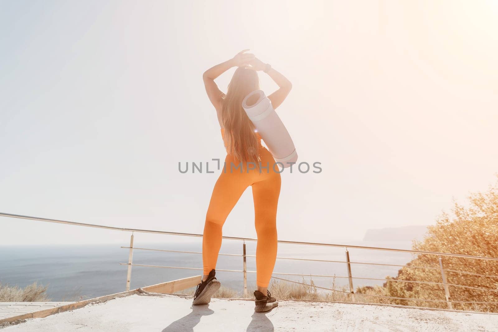 Fitness woman sea. Happy middle aged woman in orange sportswear exercises morning outdoors on yoga mat with laptop in park over ocean beach. Female fitness pilates yoga routine. Healthy lifestyle. by panophotograph