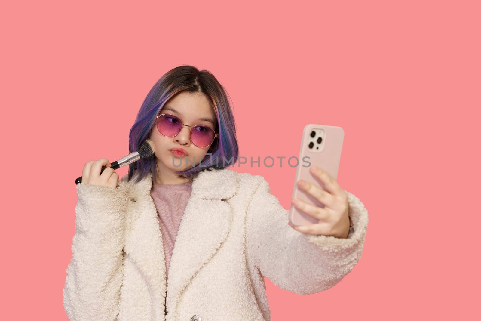 Stylish Teenager Girl, In Modern Pink Glasses, Uses Mobile Phone As Mirror While Applying Makeup With Brush Against Pink Background. Fashionable And Trendy Teenager Embracing Contemporary Beauty Practices.High quality photo