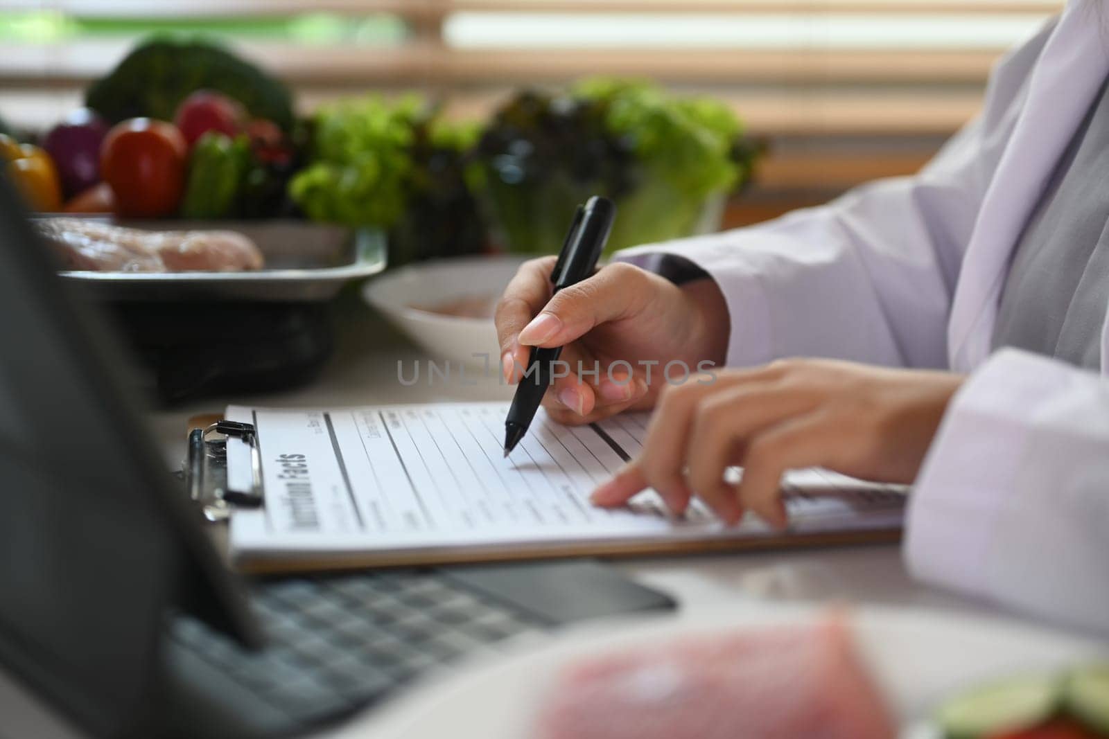 Side view of nutritionist writing diet plan and using laptop at desk with different healthy products.