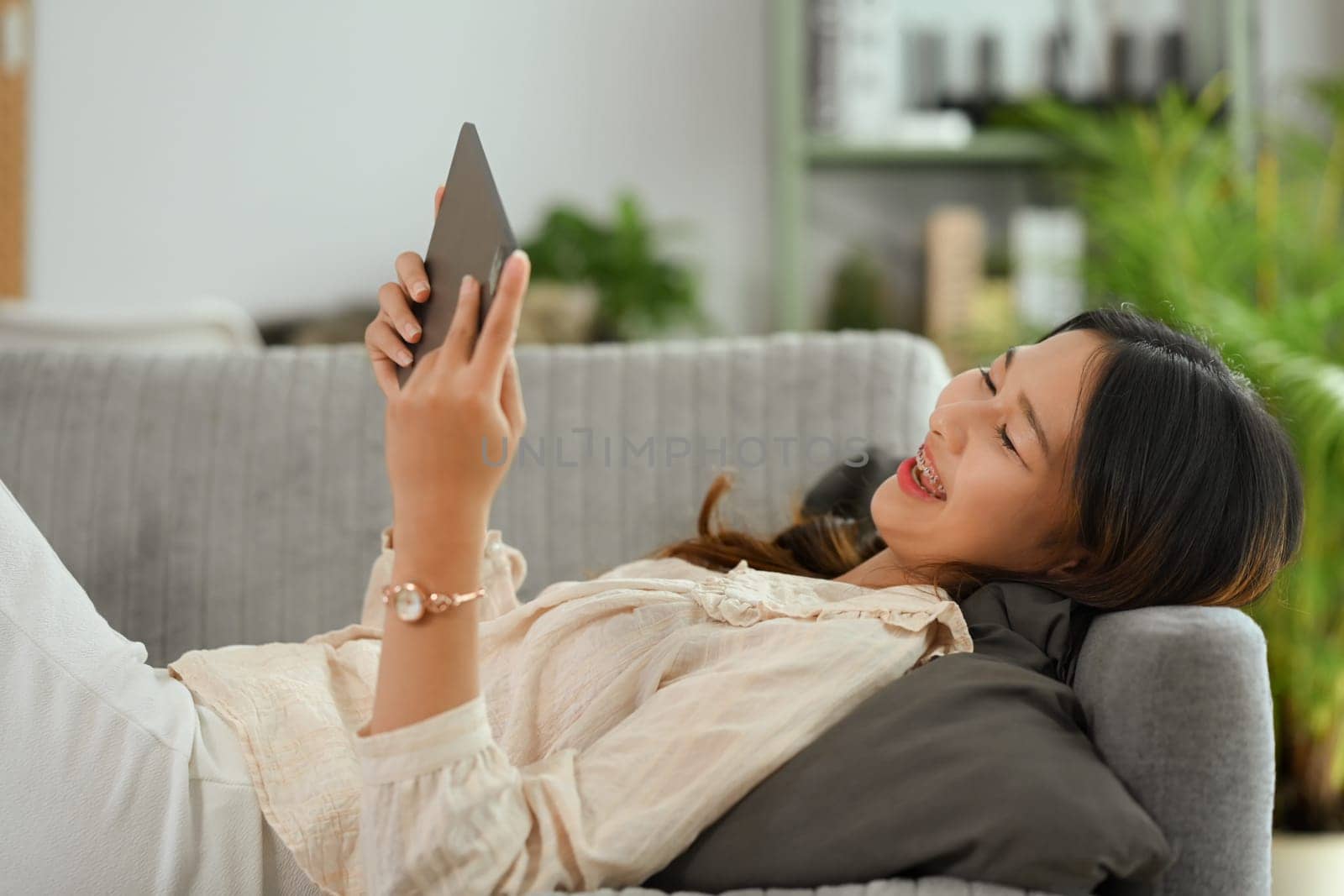 Happy young woman relaxing on couch with tablet, watching video or shopping online at home by prathanchorruangsak