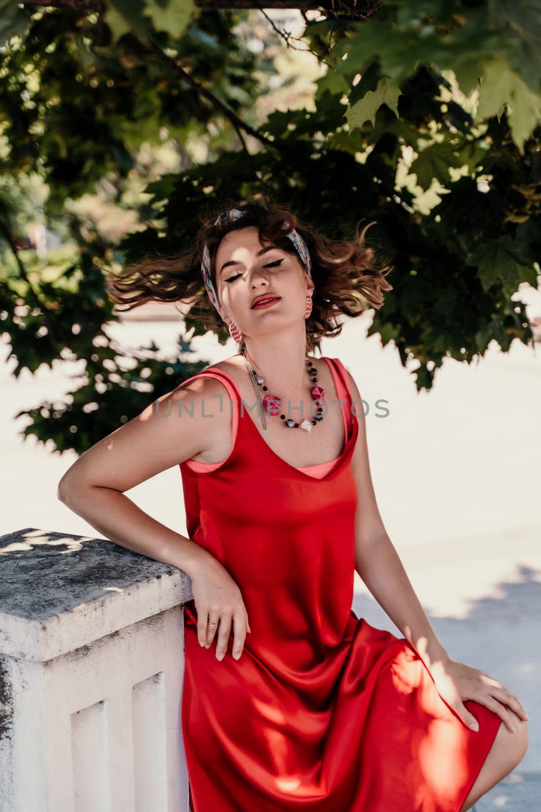 woman in a red silk dress and a bandage on her head smiles against the background of the leaves of a tree. She is leaning on the coop and looking into the camera. Vertical photo