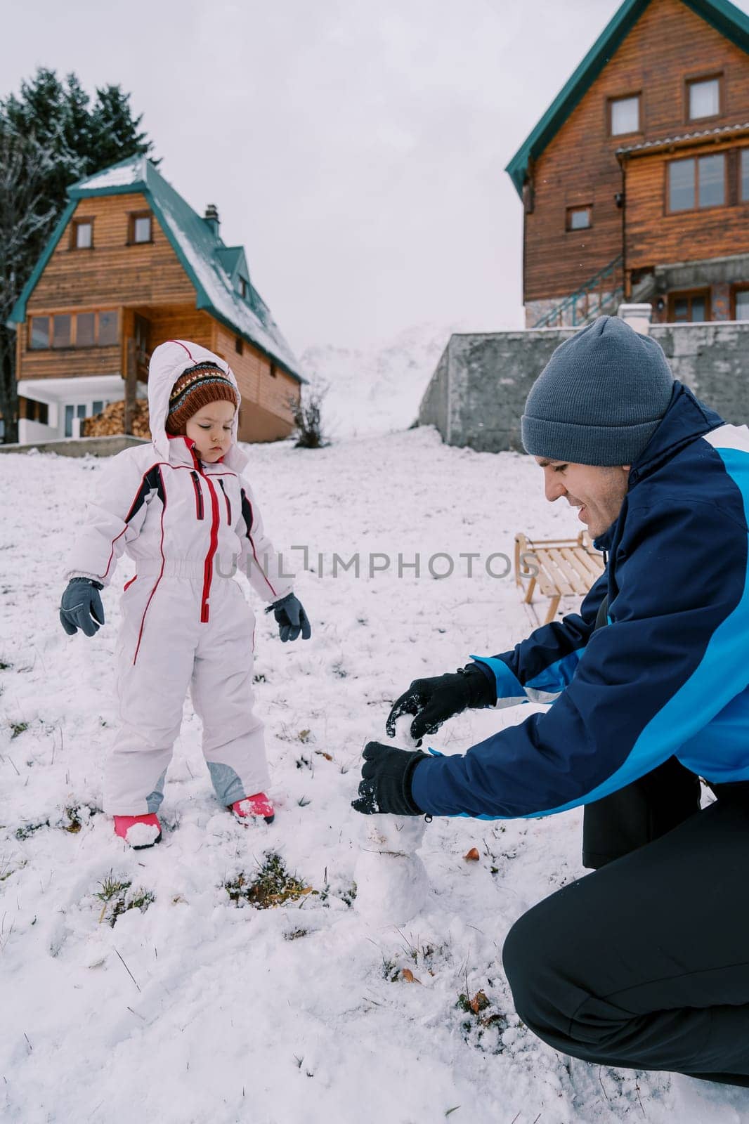 Little girl looks at her dad making a little snowman in the yard of a wooden cottage by Nadtochiy
