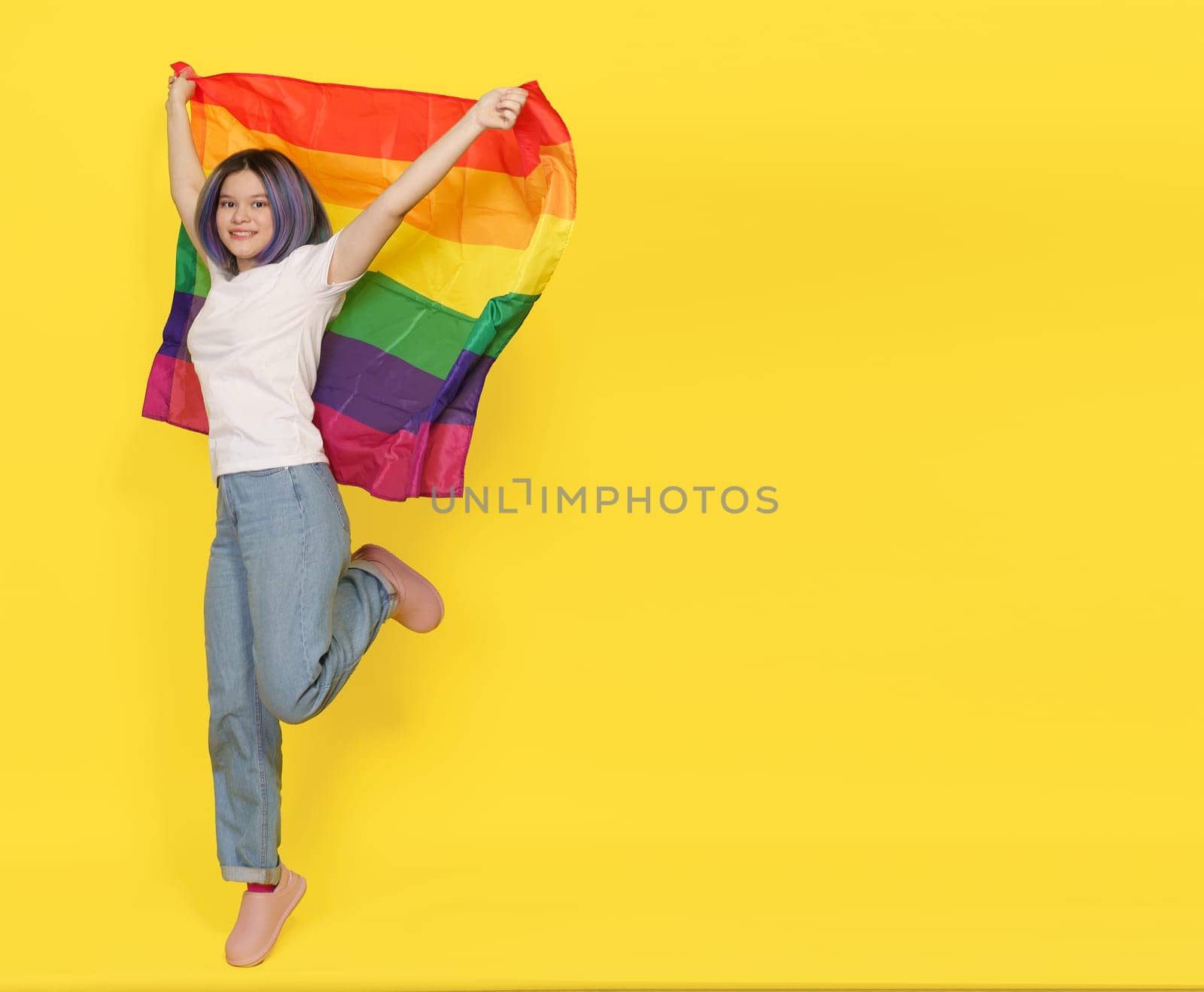 Sense Of Freedom, Equality, and LGBTQ Pride Right Of Any Person To Choose Sexual Orientation. Happy And Smiling Girl Jumps With Rainbow Flag. Isolated On Yellow Background With Copy Space by LipikStockMedia