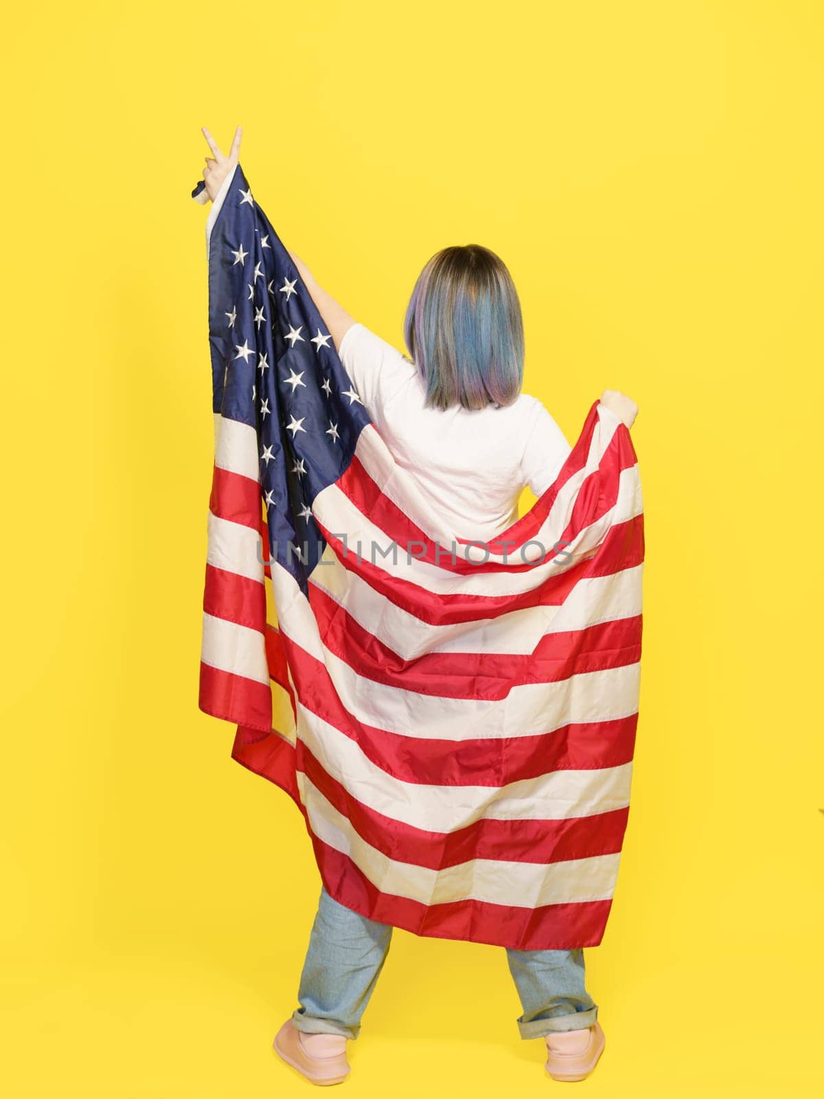 Victory Sign By Girl Covered With American Flag, Patriotic Pride, Cultural Diversity, And Freedom. Teenager Isolated On Yellow Backdrop With Copy Space For Your Text. Full-Height Backside View by LipikStockMedia