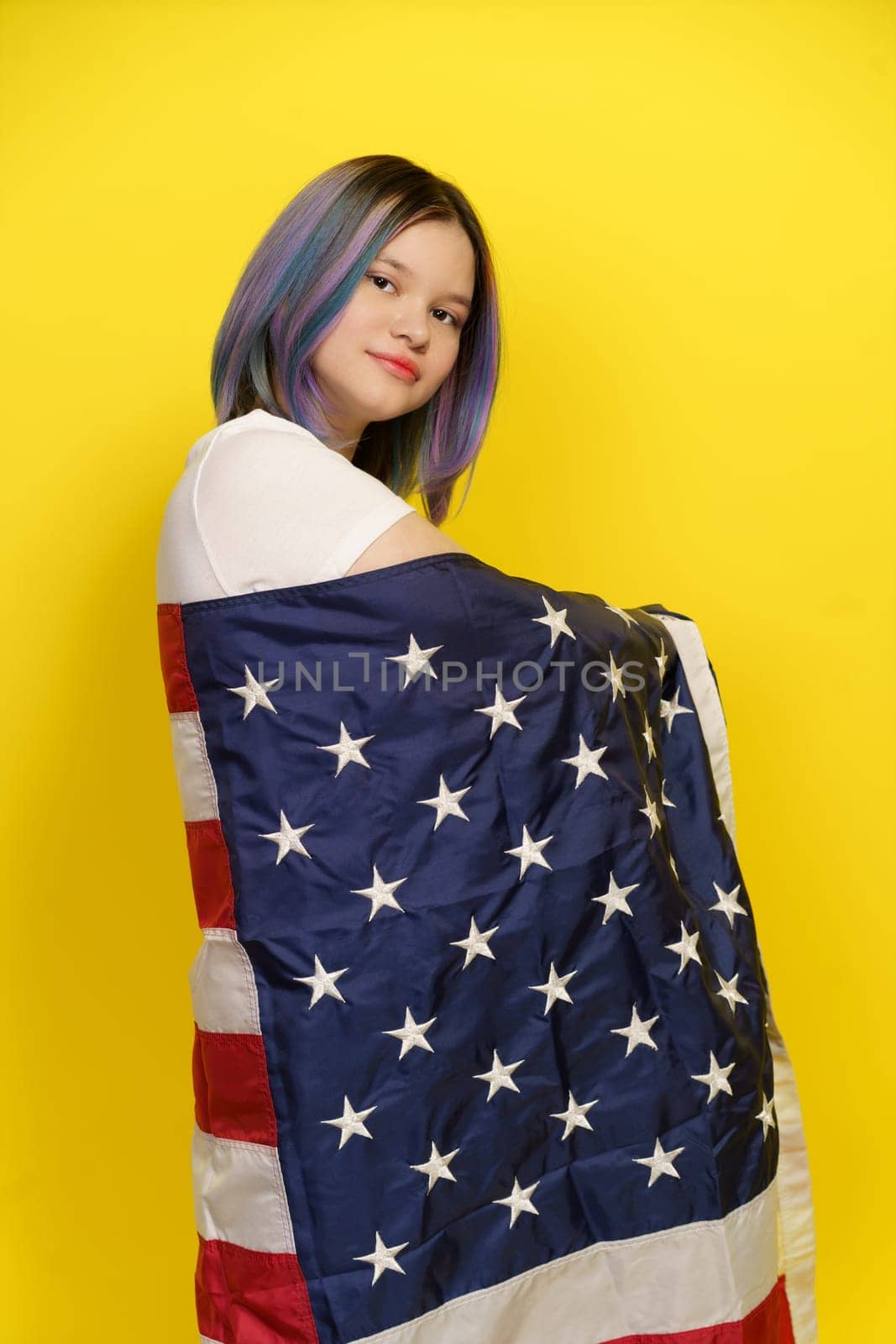 Proud Citizen With Multicolored Hair Poses For Camera In Studio, Wrapped In American Flag On Yellow Background. Patriotism, Pride, And Celebration Of Cultural Diversity. Proud American Teenager. High quality photo