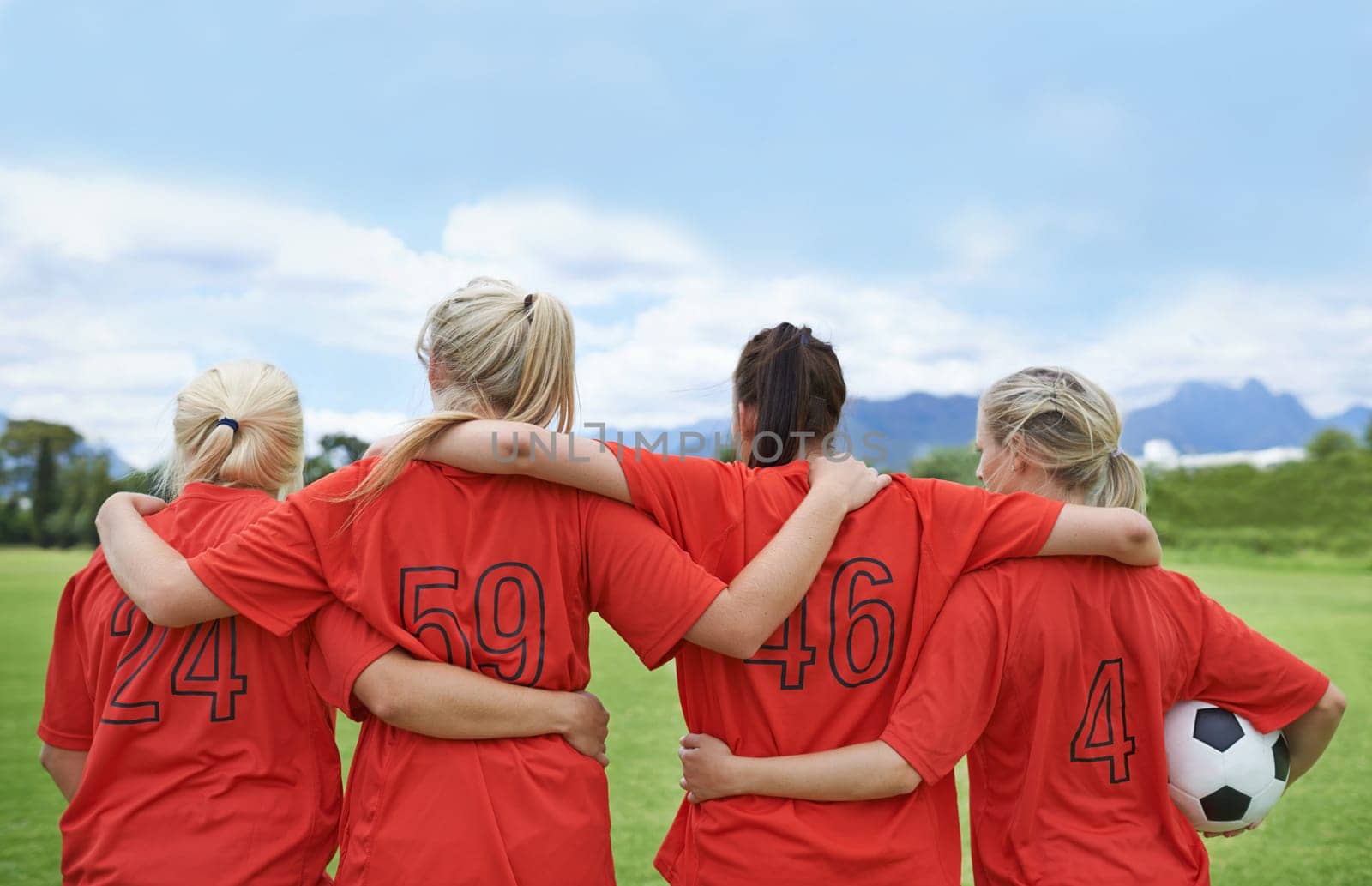 Woman, back and hug in soccer of sports team ready for match, game or outdoor practice with blue sky. Rear view of female person or group of football players standing in unity with ball in nature.