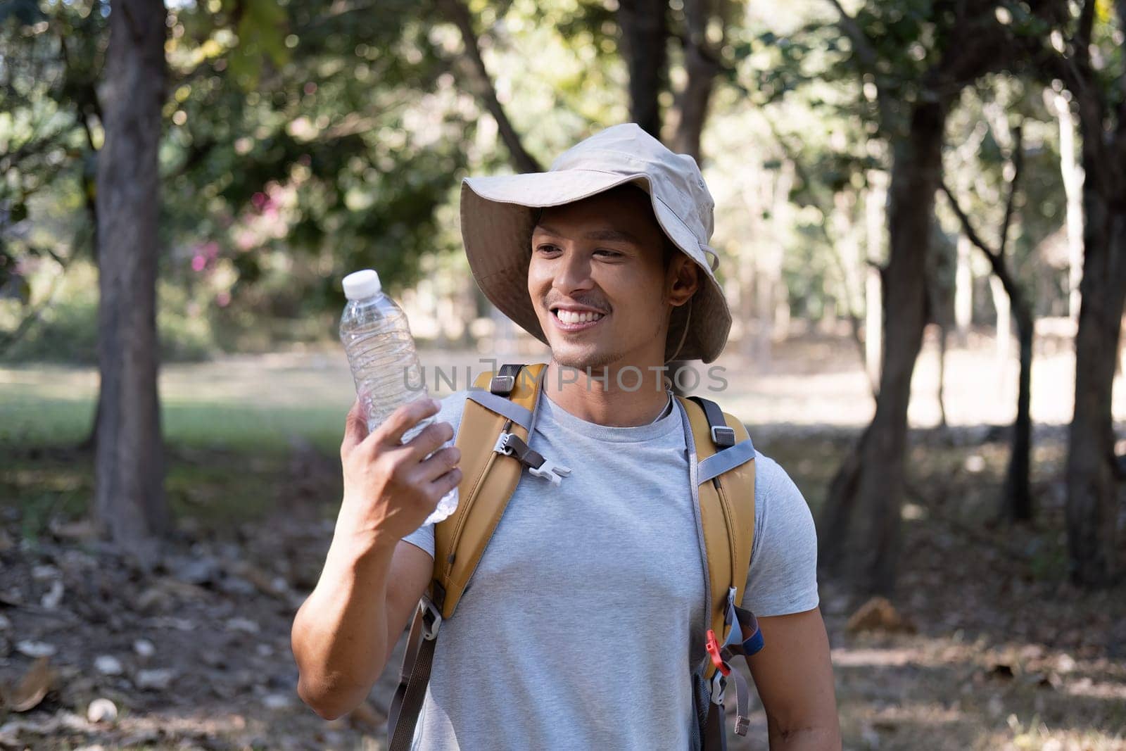 Asian male traveler carrying a large backpack drinks water from a bottle while resting during a hike..