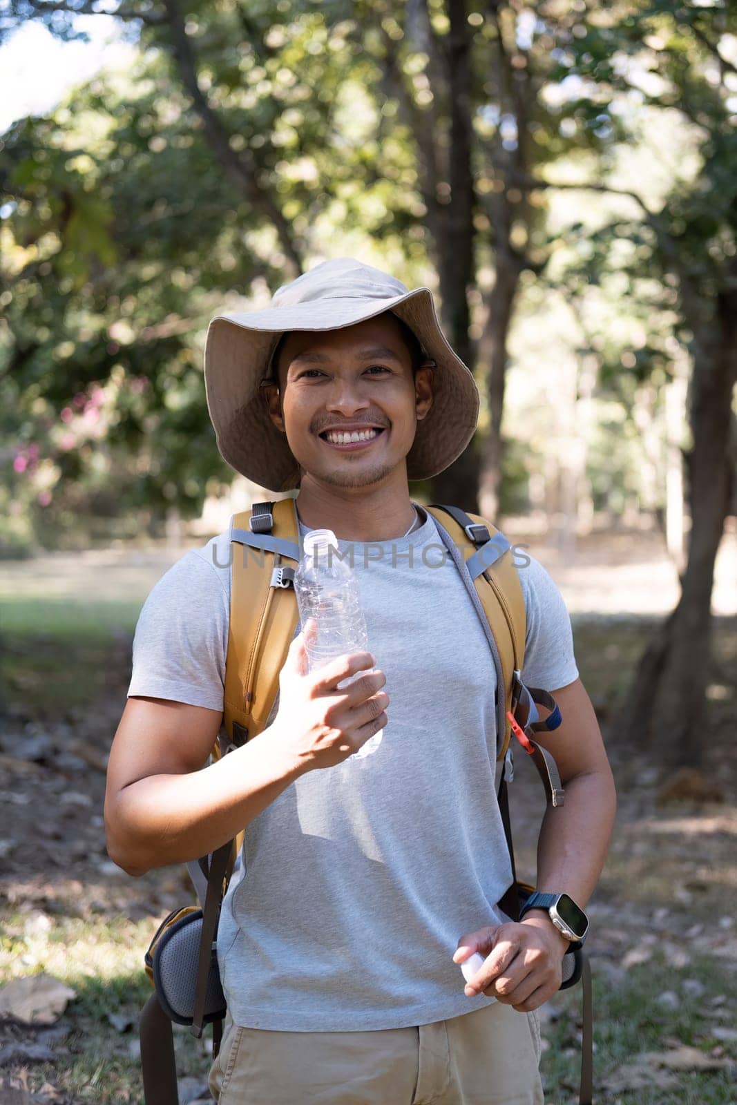 Asian male traveler carrying a large backpack drinks water from a bottle while resting during a hike..