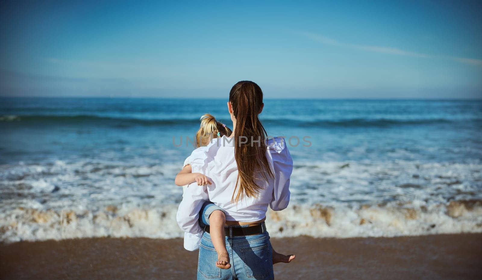 Rear view of a young woman, loving affectionate mother carrying little daughter, enjoying a weekend together, walking along a tropical beach. Waves splashing on the background. People and Nature