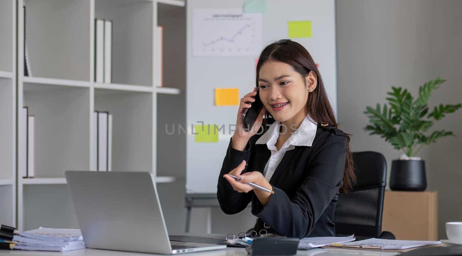 Cheerful Asian businesswoman talking on the phone working in modern office Happy Asian businesswoman company manager wearing a suit is talking on the phone sitting in the office. by wichayada