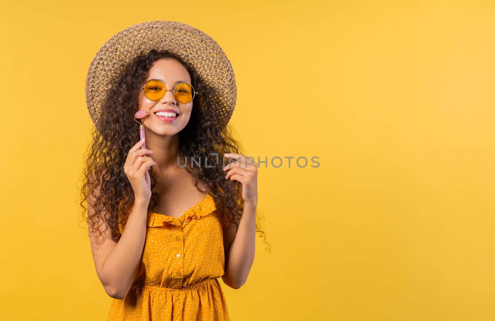 Young woman doing face massage with rose quartz stone roller, yellow background by kristina_kokhanova