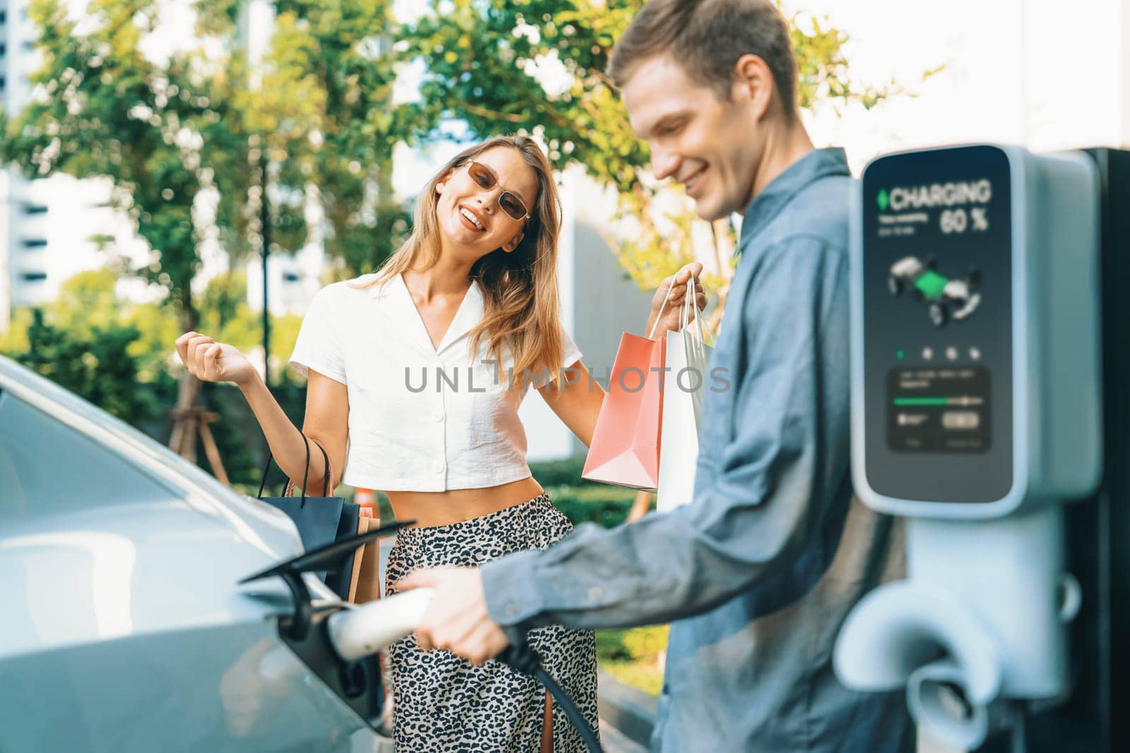 Young couple travel with EV electric car charging in green sustainable city outdoor garden in summer shows urban sustainability lifestyle by green clean rechargeable energy of electric vehicle innards