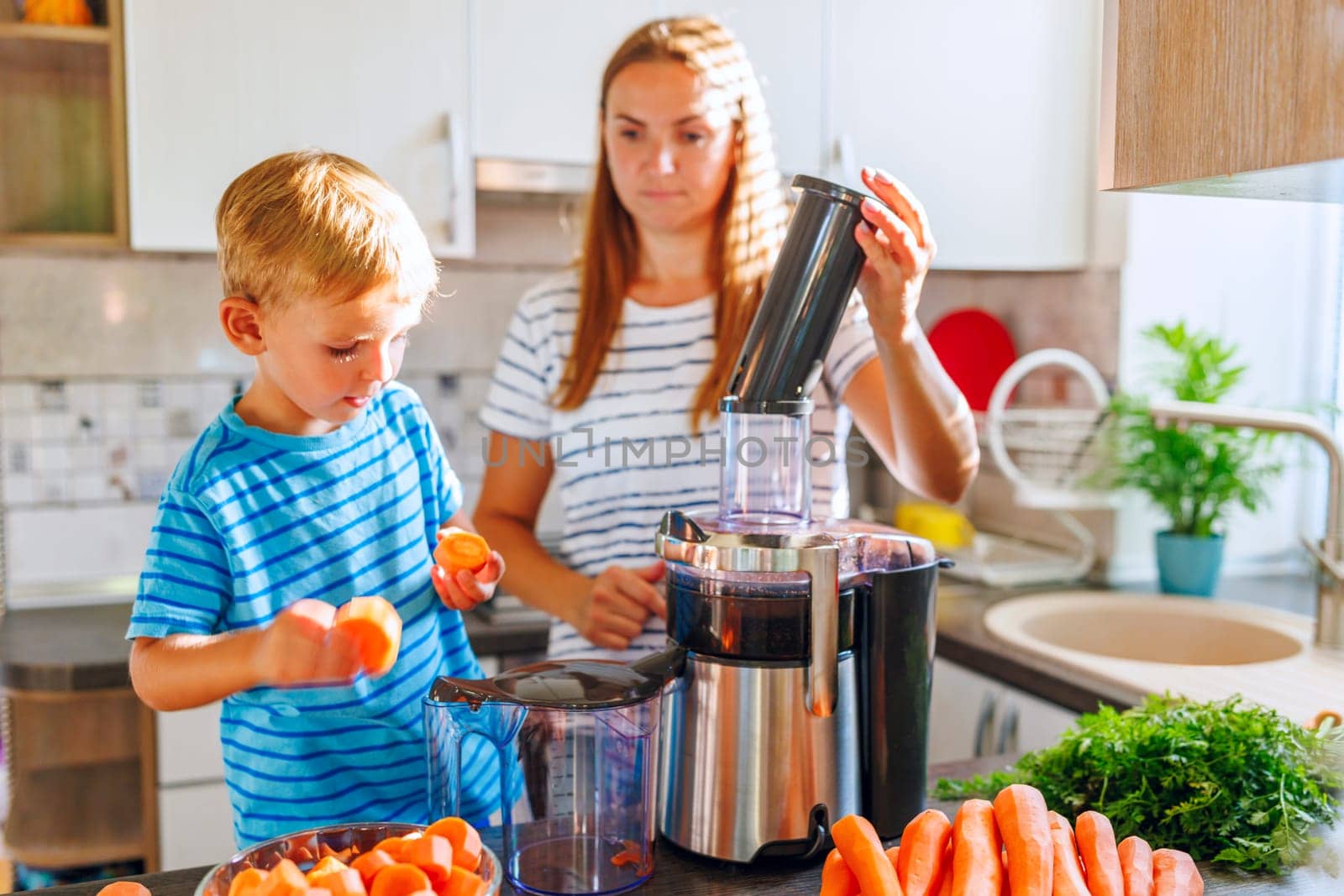 In a bright kitchen, a mother and her young son prepare healthy carrot juice using a juicer, enjoying a moment of family bonding.