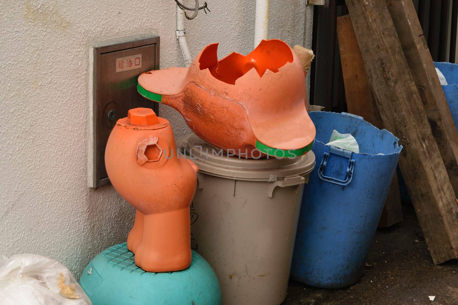 A broken plastic statue pots on a trash bin with other waste containers nearby, depicting waste and recycling.