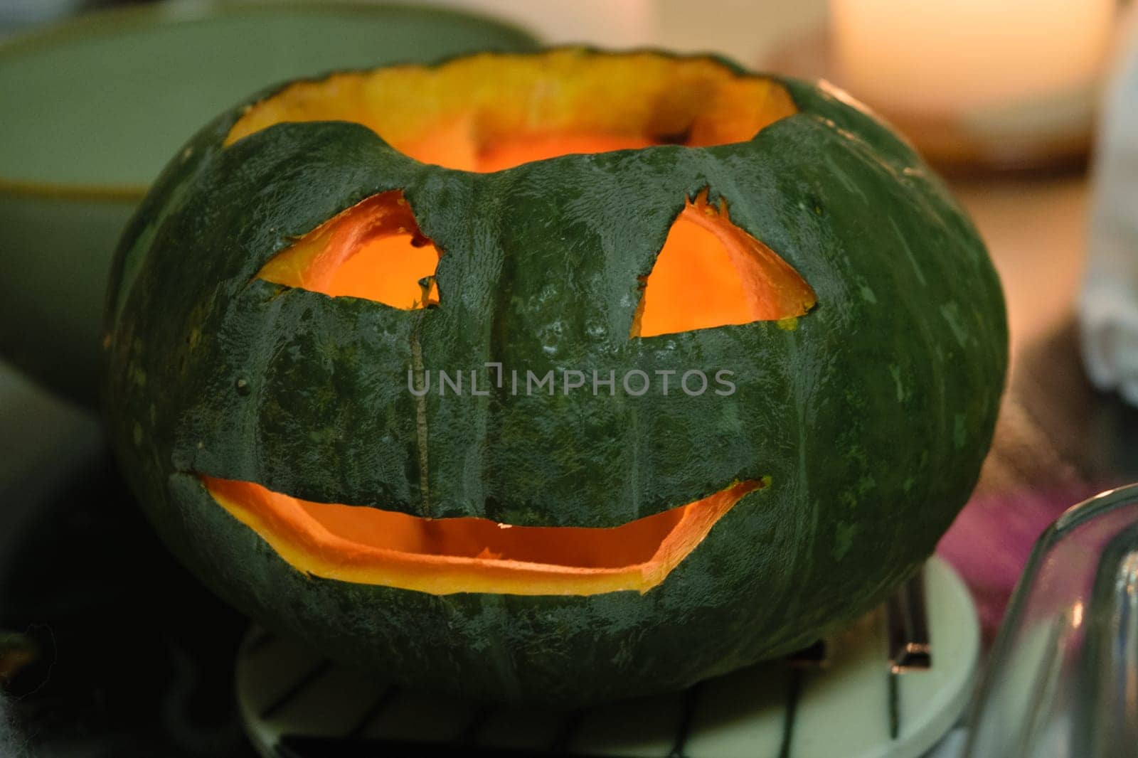 Hand-carved green pumpkin with a glowing interior and a smiling jack-o'-lantern face, set against a soft-focus background.