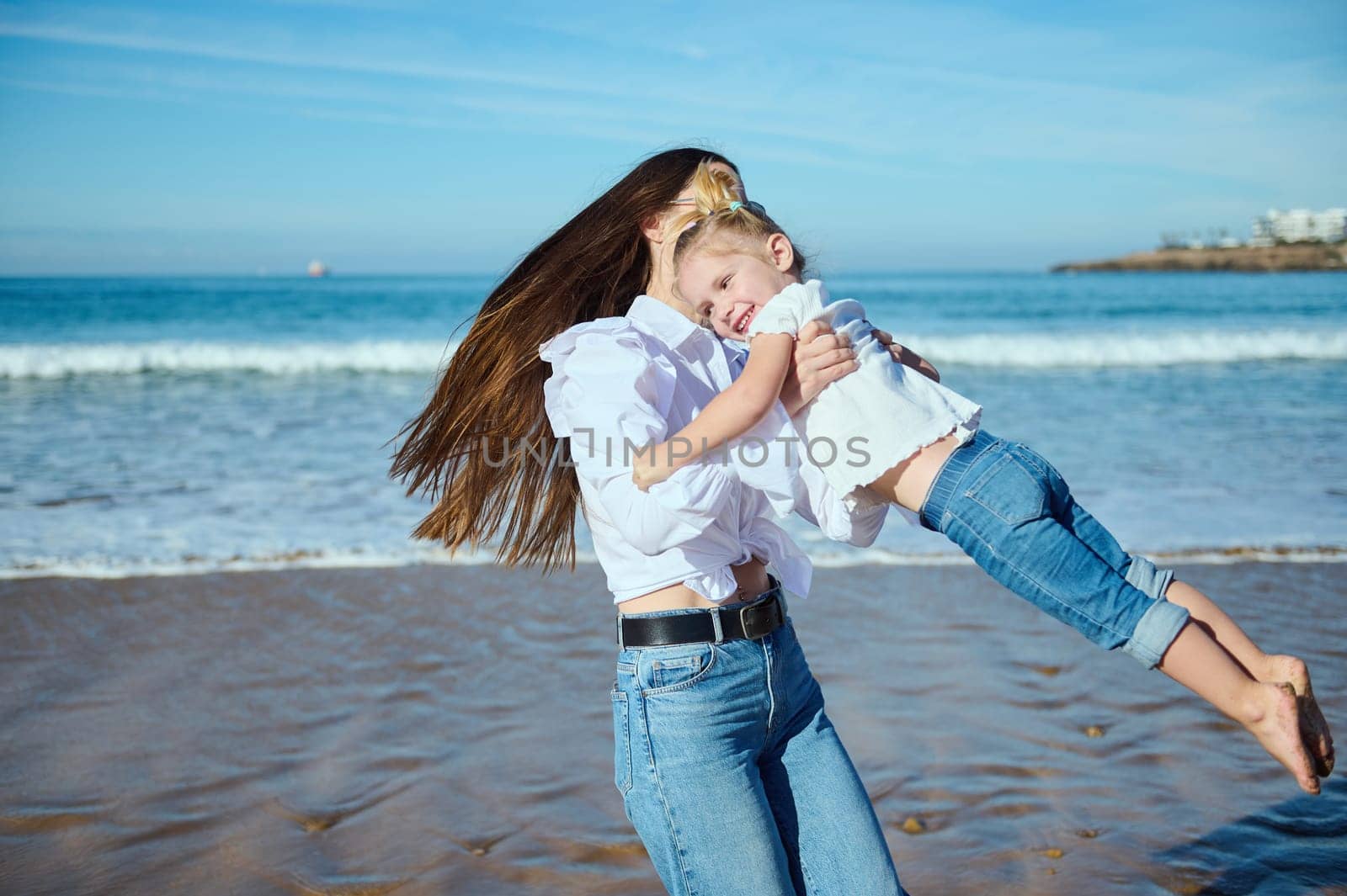 Happy young mother plays with her cute kid on the beach, laughing and turning around, leaving footstep on the wet sand by artgf