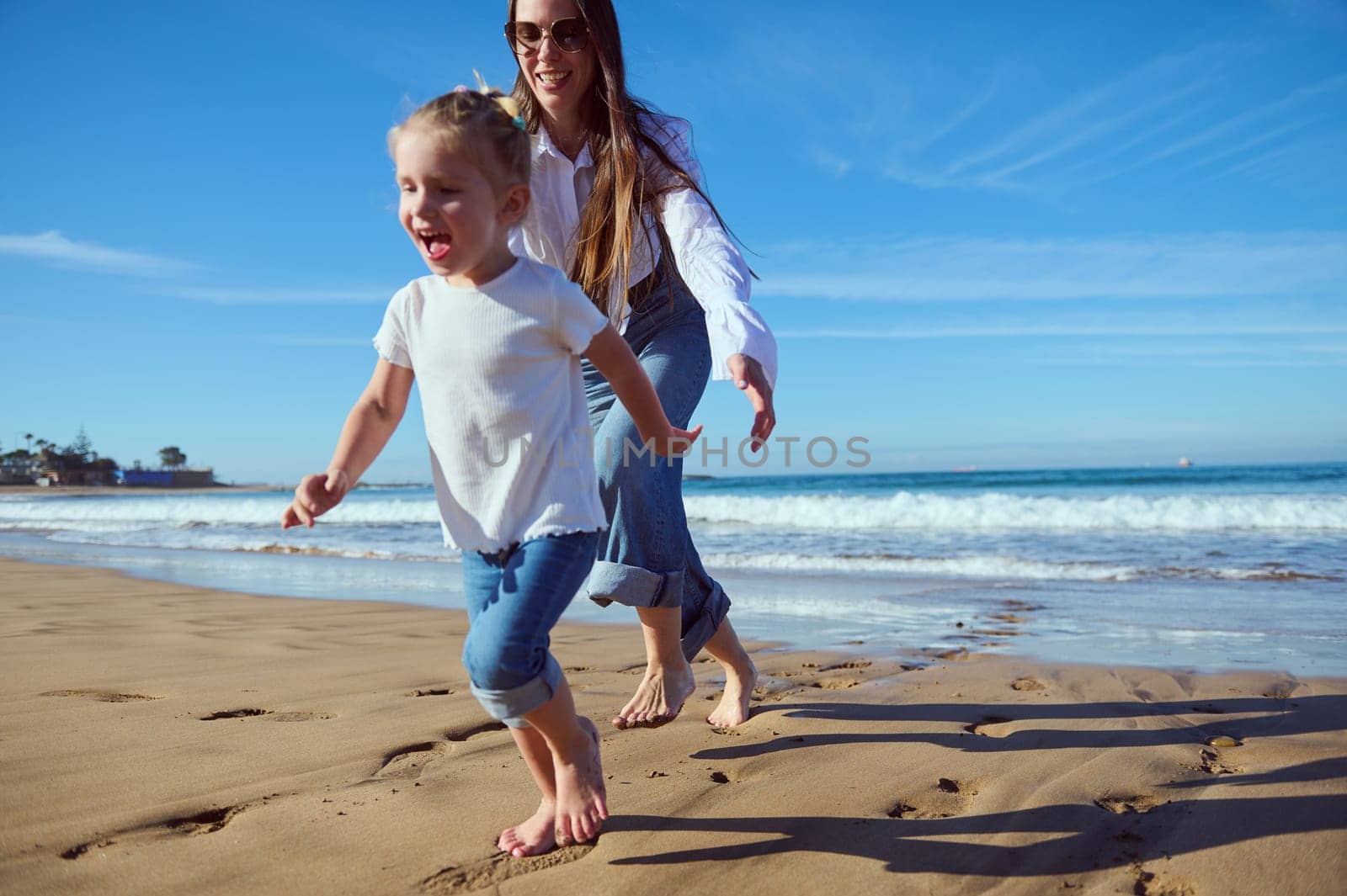 Beautiful active little child girl and her mother playing together and running barefoot on the sandy beach by artgf