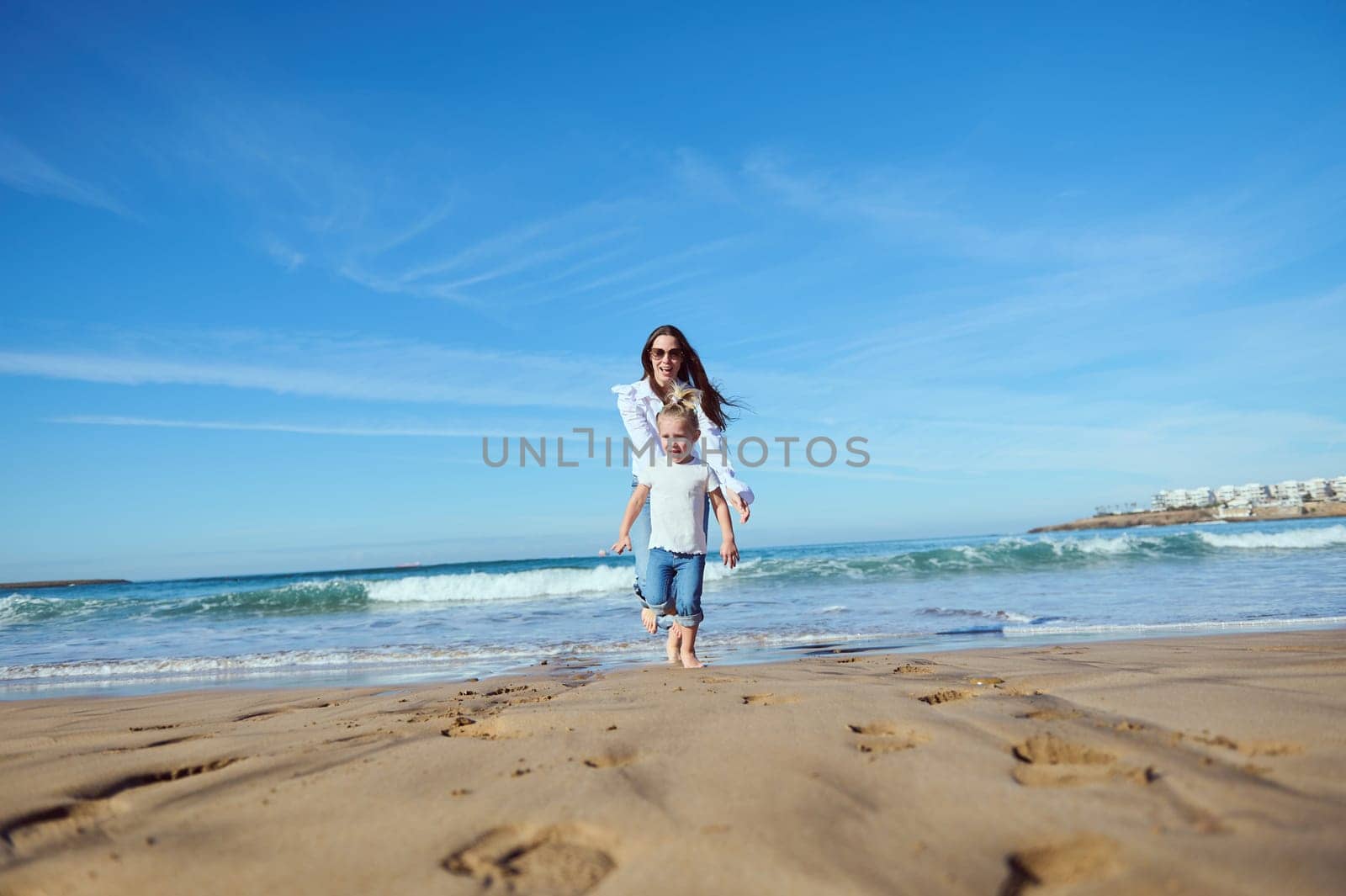 Cheerful happy mom catches her daughter while running barefoot, playing together on beach, leaving footsteps on wet sand by artgf