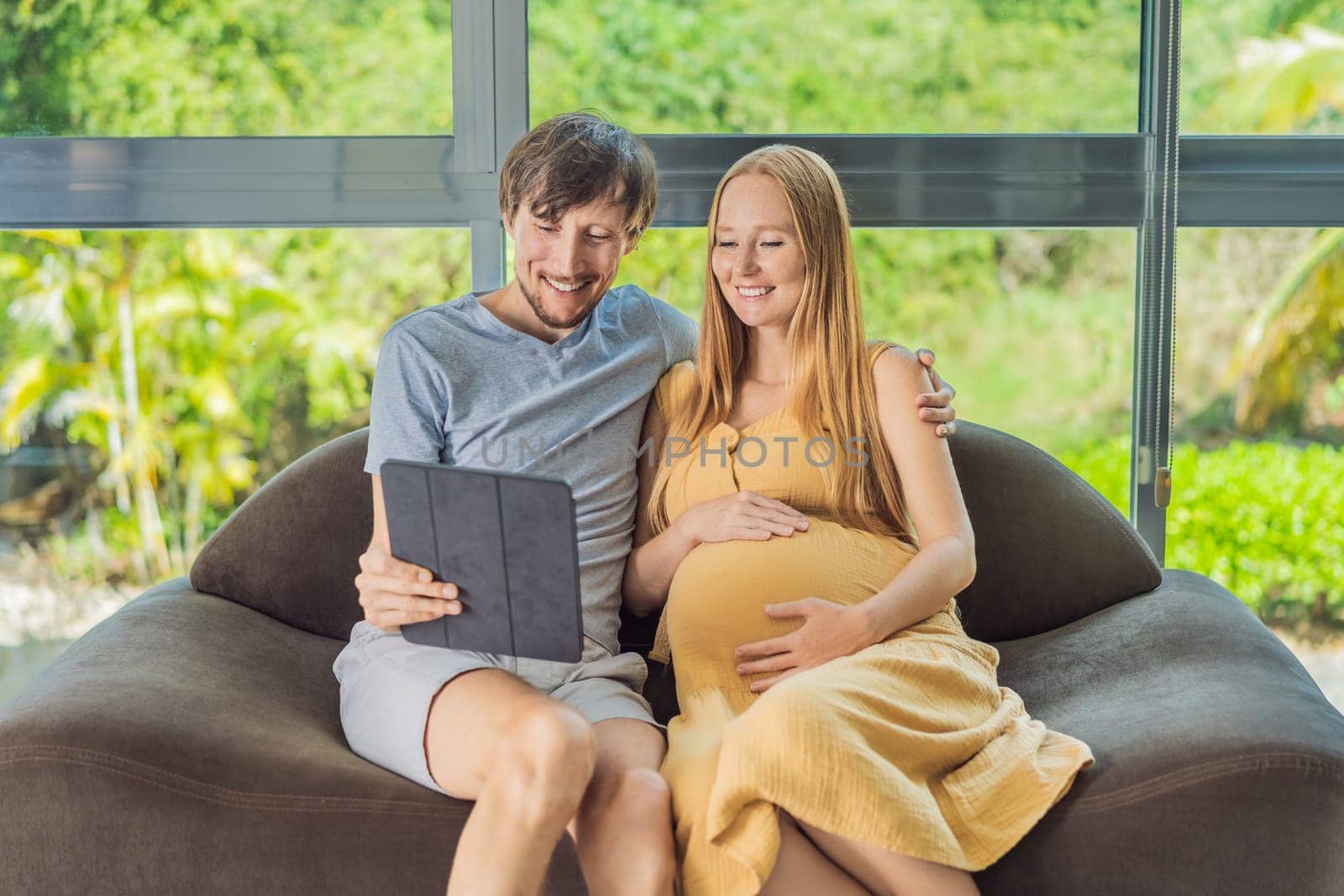 A smiling husband and his pregnant wife share a moment of joy, gazing at a tablet screen, their faces glowing with anticipation and happiness.