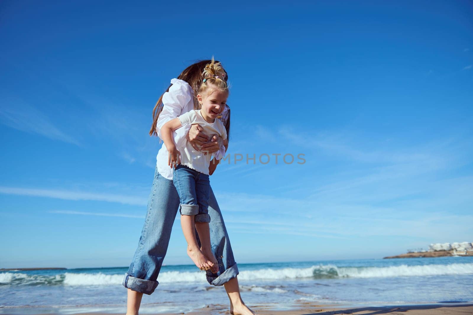 Mother and daughter playing together on the beach. Happy little kid girl smiling and expressing positive emotions while her mom carrying her, playing with warm waves