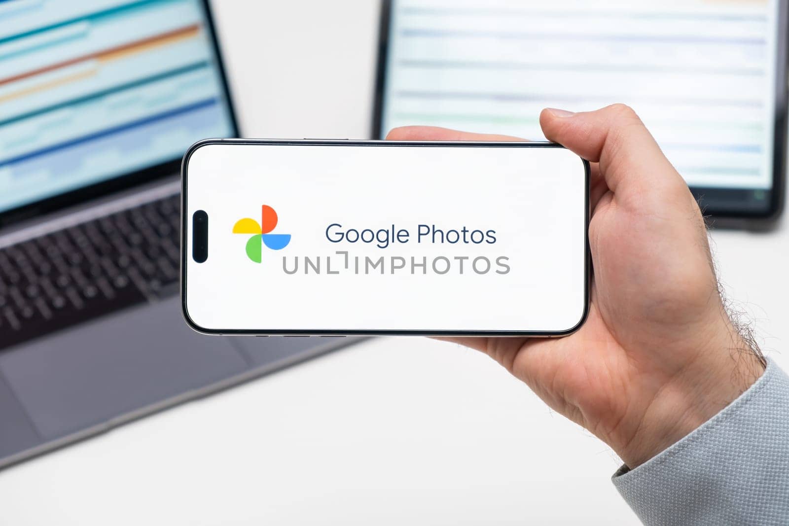 Google Photos logo of app on the screen of mobile phone held by man in front of the laptop and tablet by vladimka