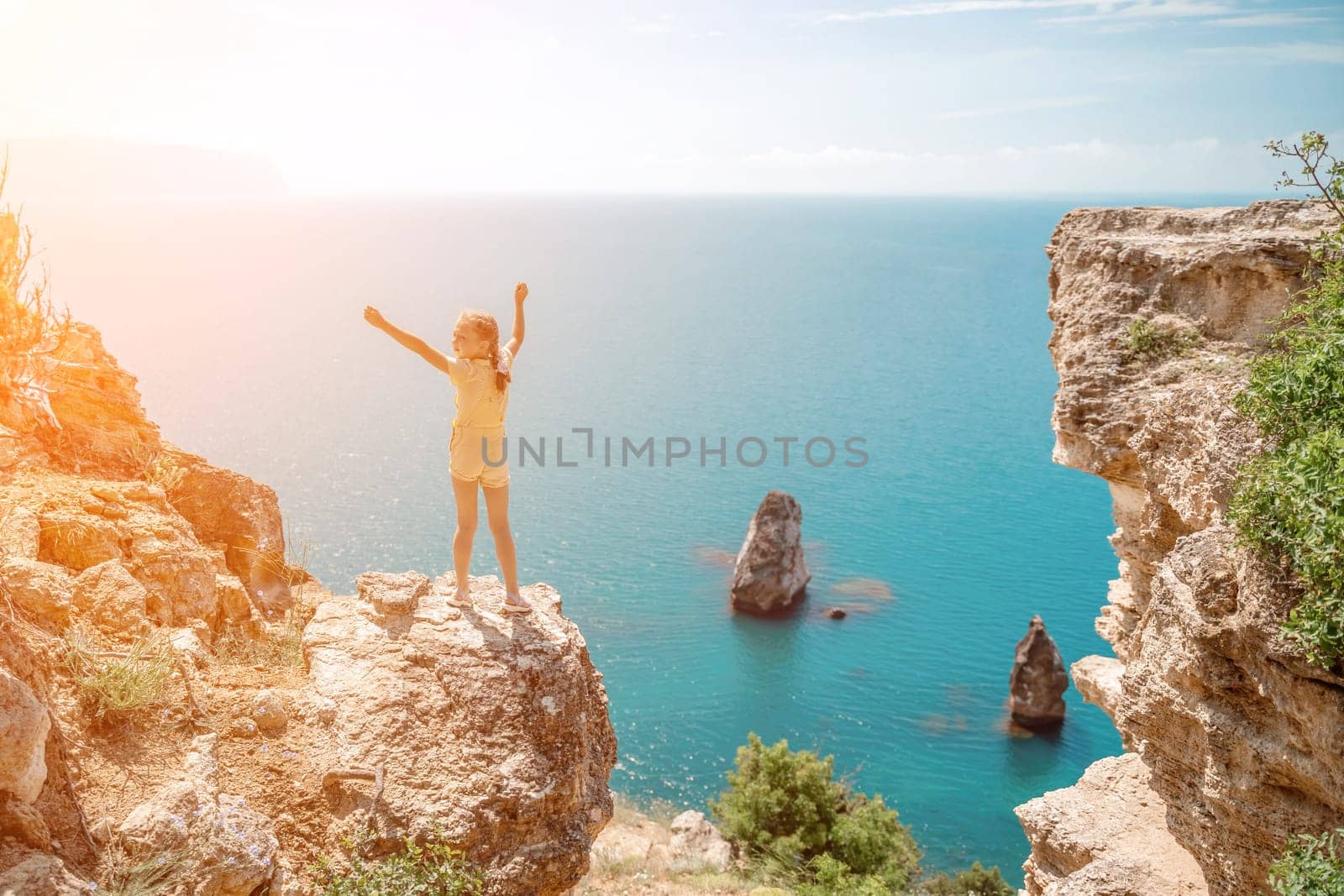 Happy girl stands on a rock high above the sea, wearing a yellow jumpsuit and sporting braided hair, depicting the idea of a summer vacation by the sea. by Matiunina