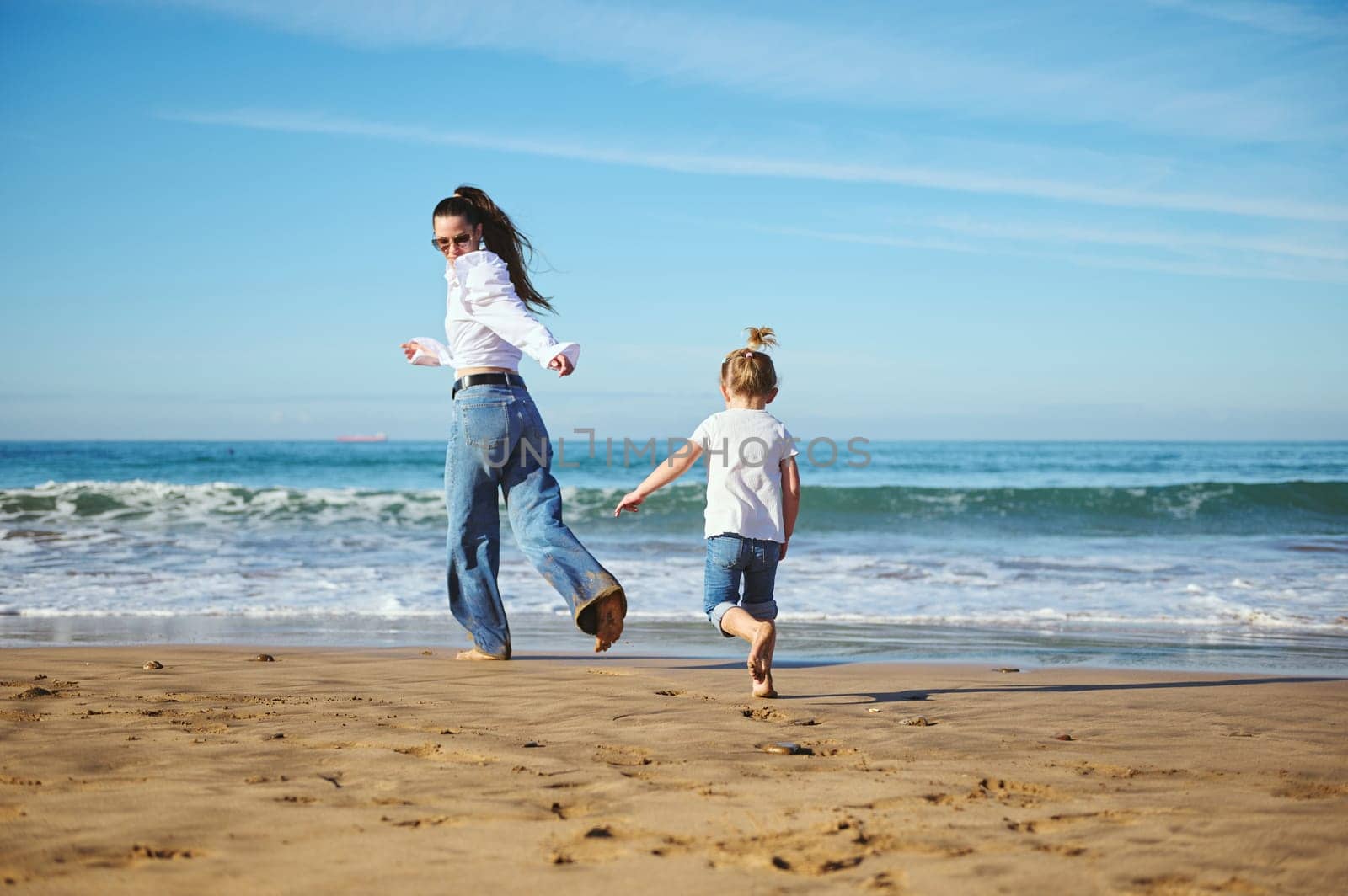 Full length portrait of an authentic family of a young mother and cute daughter, running barefoot on the beach, leaving footsteps on the wet sand while beautiful waves splashing and washing their feet