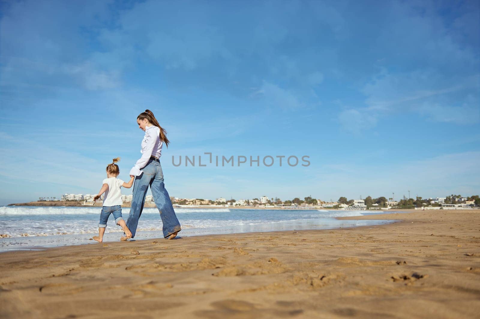 Young woman, mother and her daughter walking barefoot on warm water on waves, playing together, spending happy nice time