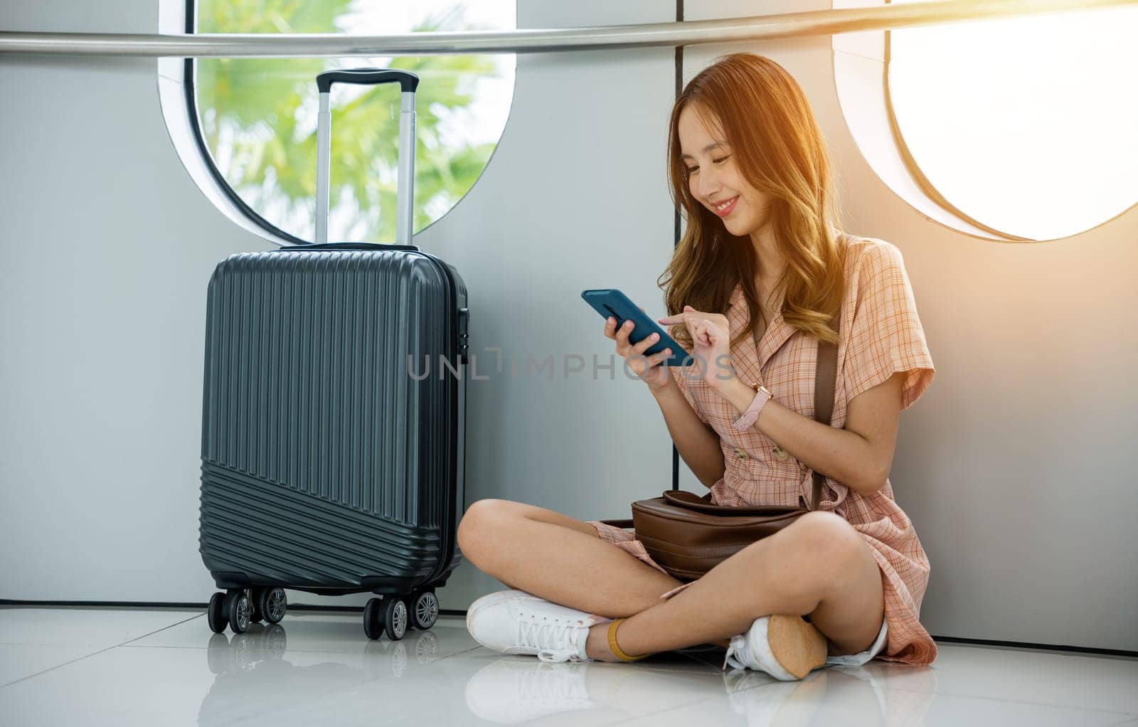 Young woman sitting next to her suitcase and backpack at the airport terminal, using her phone to book her next travel destination. travel in vacation summer