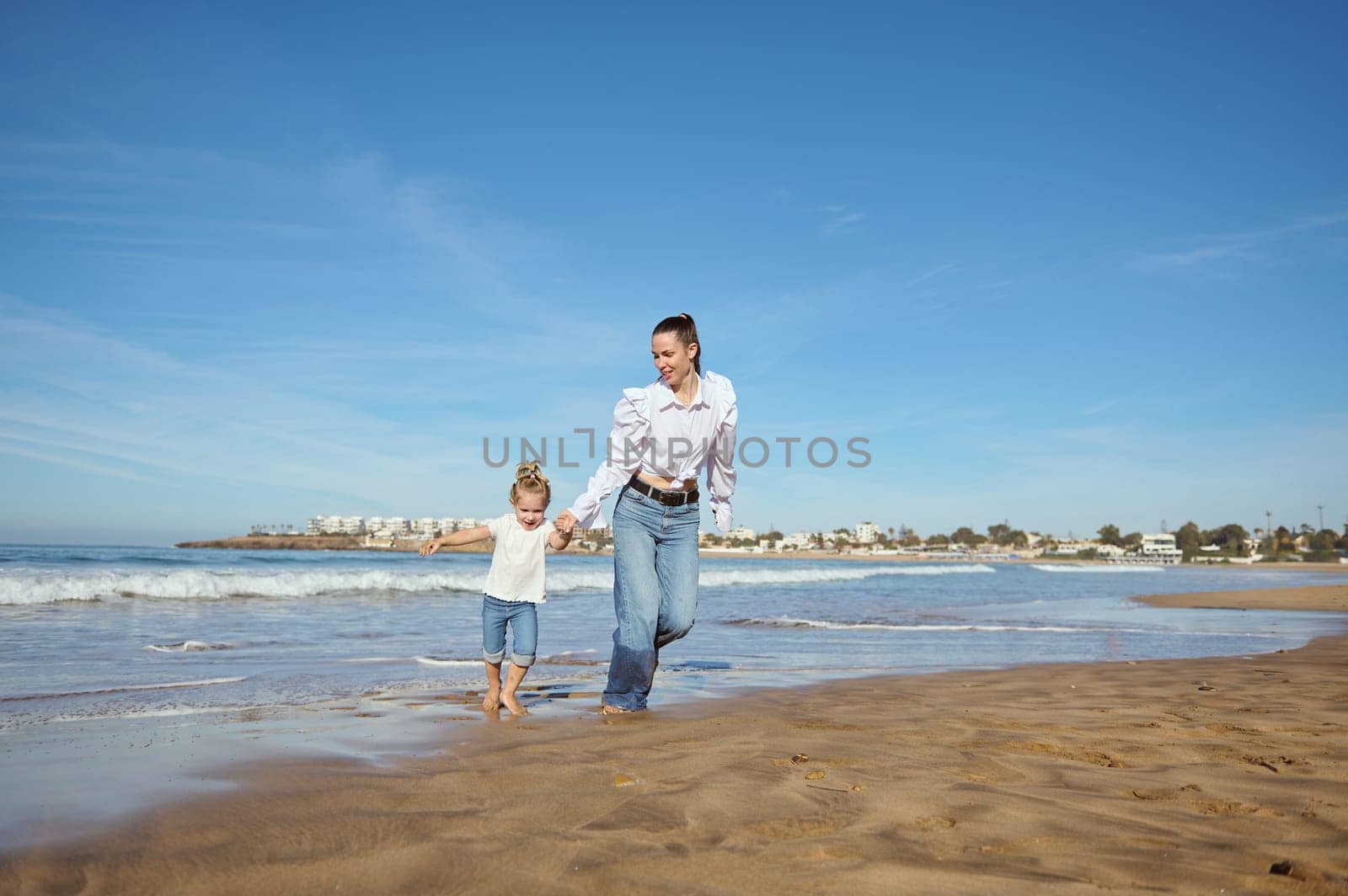 Young mother and her daughter holding hands while running barefoot on warm water on waves, playing together on the beach by artgf