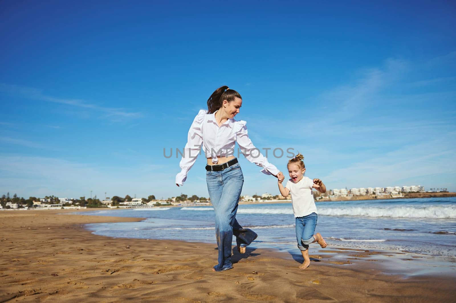 Cheerful young woman, mother and her daughter running barefoot on warm water on waves, playing together, spending happy nice time. Family relationships. People. Lifestyle. Leisure activity
