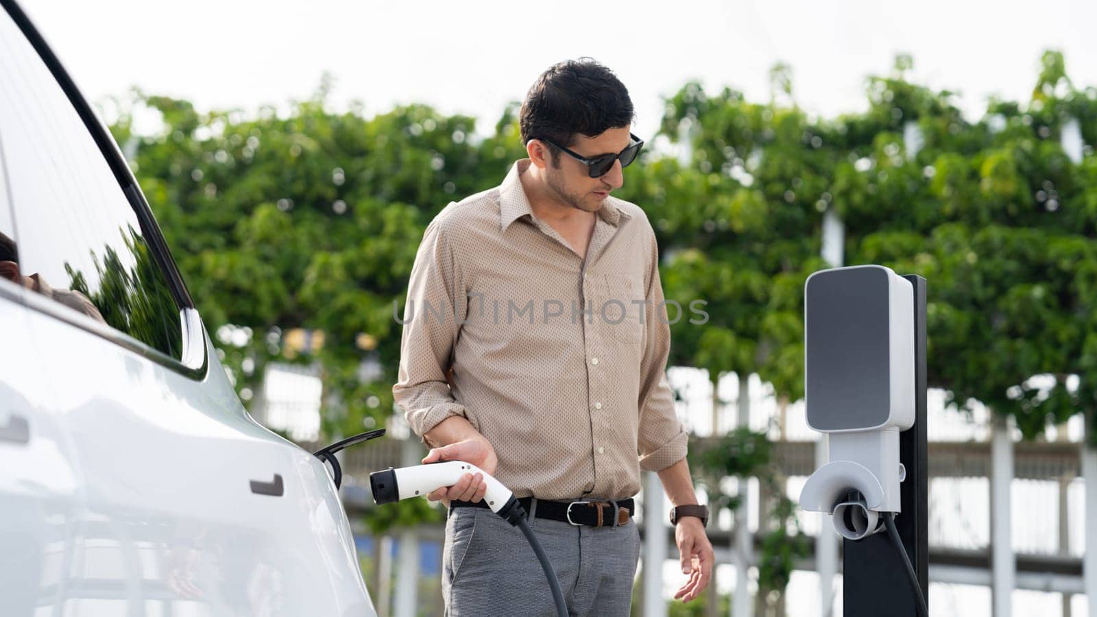 Young man recharge electric car's battery from charging station in outdoor green city park. Rechargeable EV car for sustainable environmental friendly urban travel. Panorama Expedient