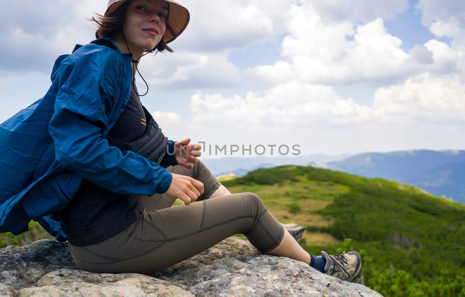 A happy young girl in a hat and windbreaker sits on a rock and enjoys an amazing view with mountains, hills and forest during her journey, hiking in the wild. A woman spends an active lifestyle.
