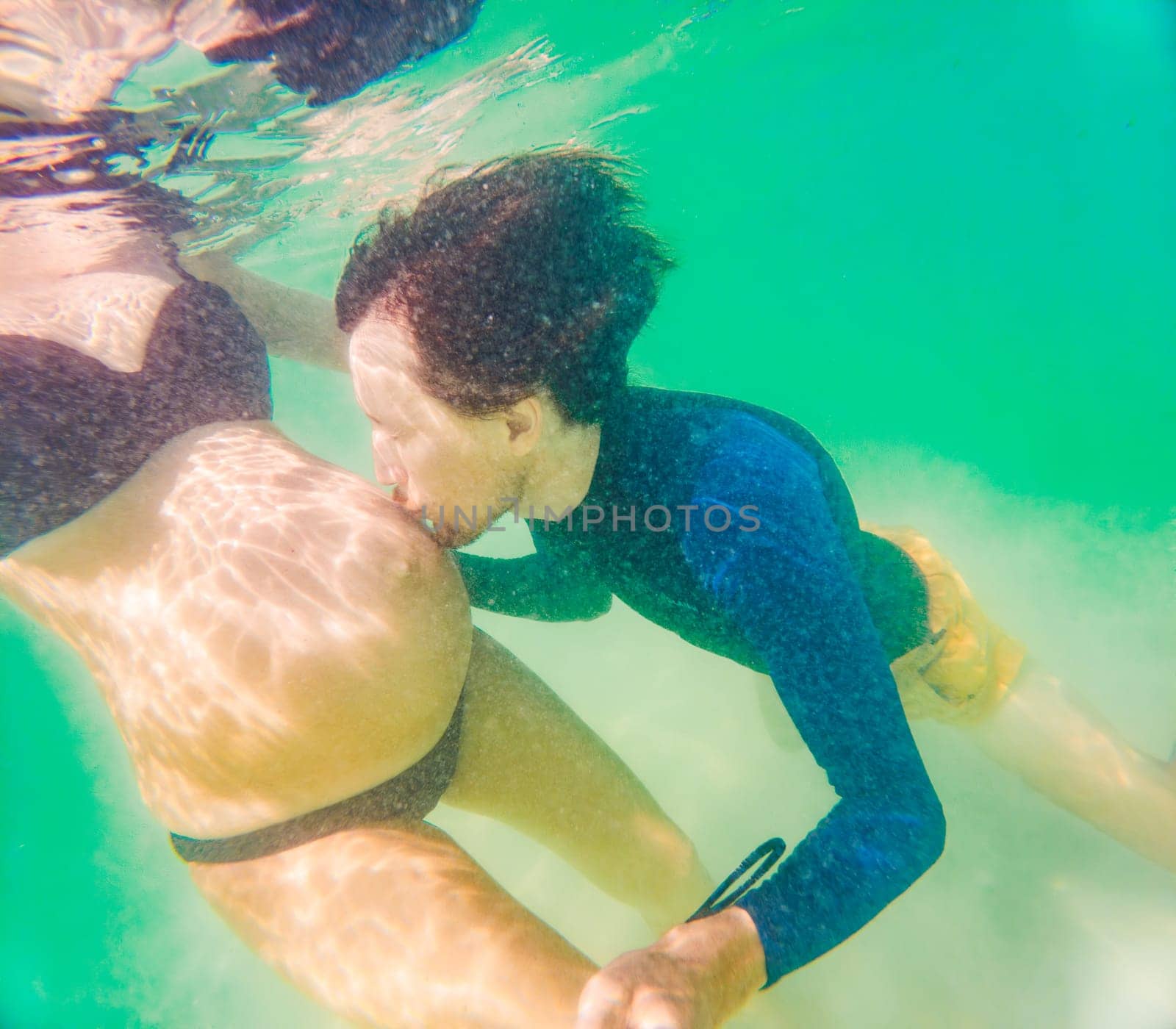 Amidst the tranquil turquoise sea, a pregnant woman and her husband share a serene moment, celebrating the beauty of impending parenthood in the gentle waves.