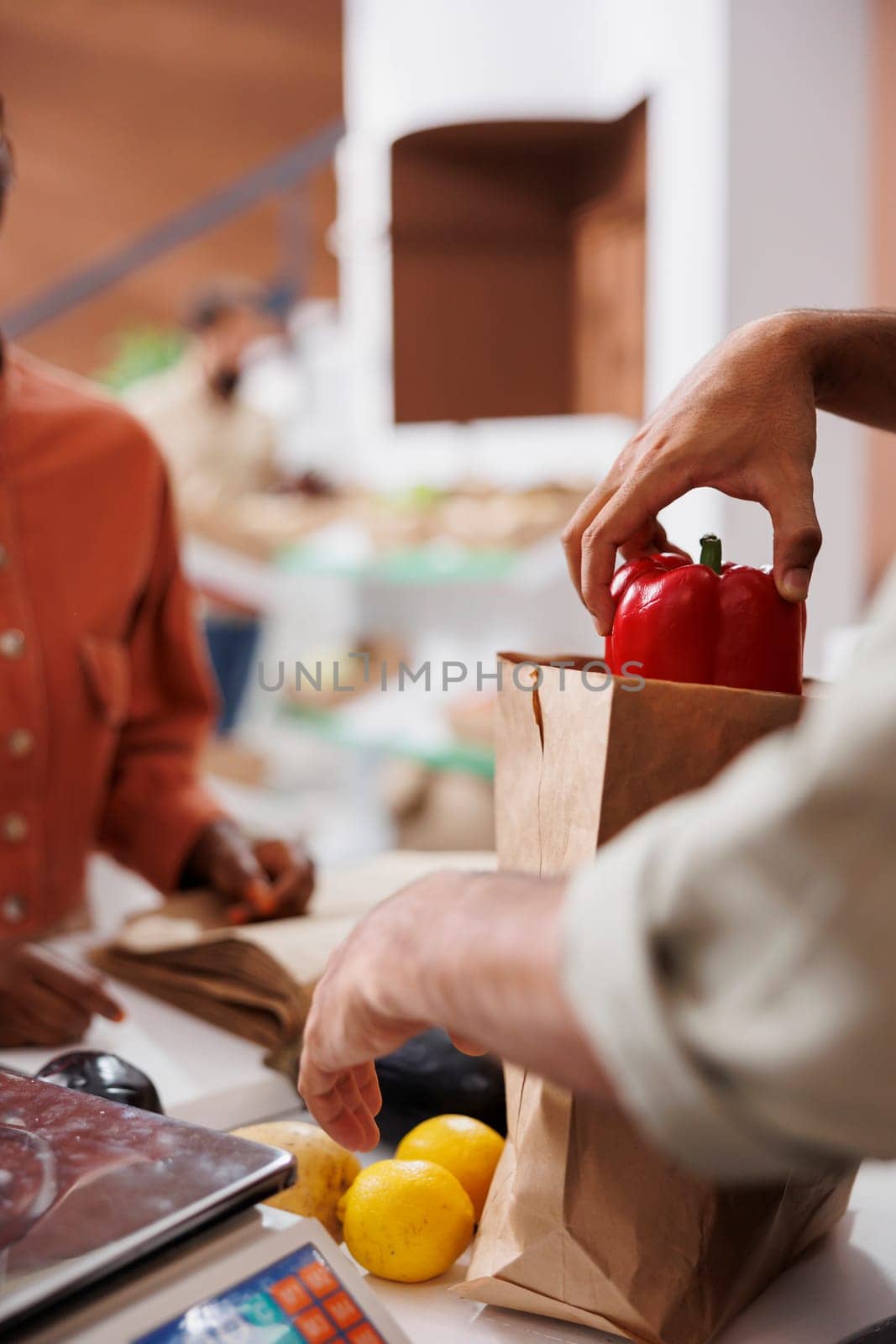 Close-up of vendor at checkout counter placing freshly harvested produce into paper bag for female customer. A person is seen in the image packing locally grown fruits and vegetables for takeaway.