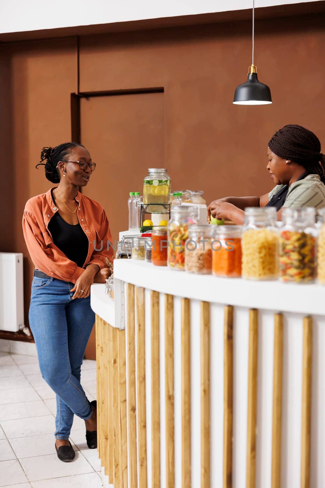 African American vendor checks out groceries at a local market offering eco-friendly, organic products. Black women at cashier desk conversing about locally grown nutritious organic products.
