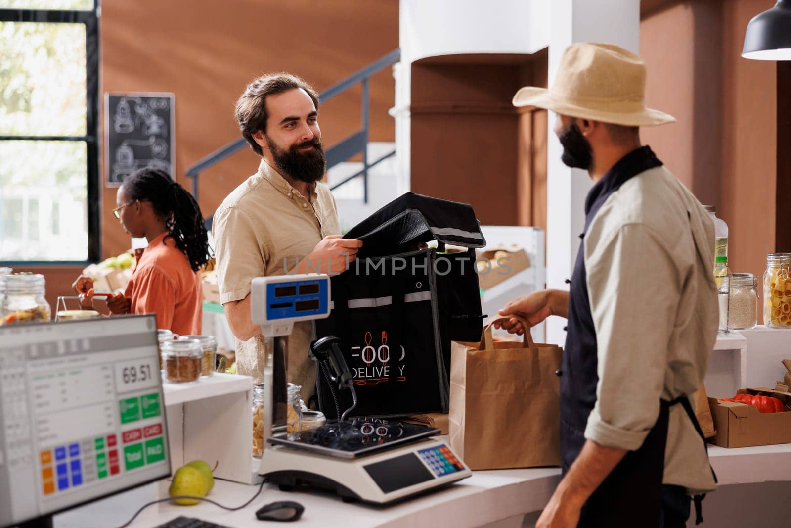 Caucasian male courier arrives at checkout counter ready to receive packaged grocery order from cashier. Food delivery man with backpack retrieves takeaway from vendor at local market.
